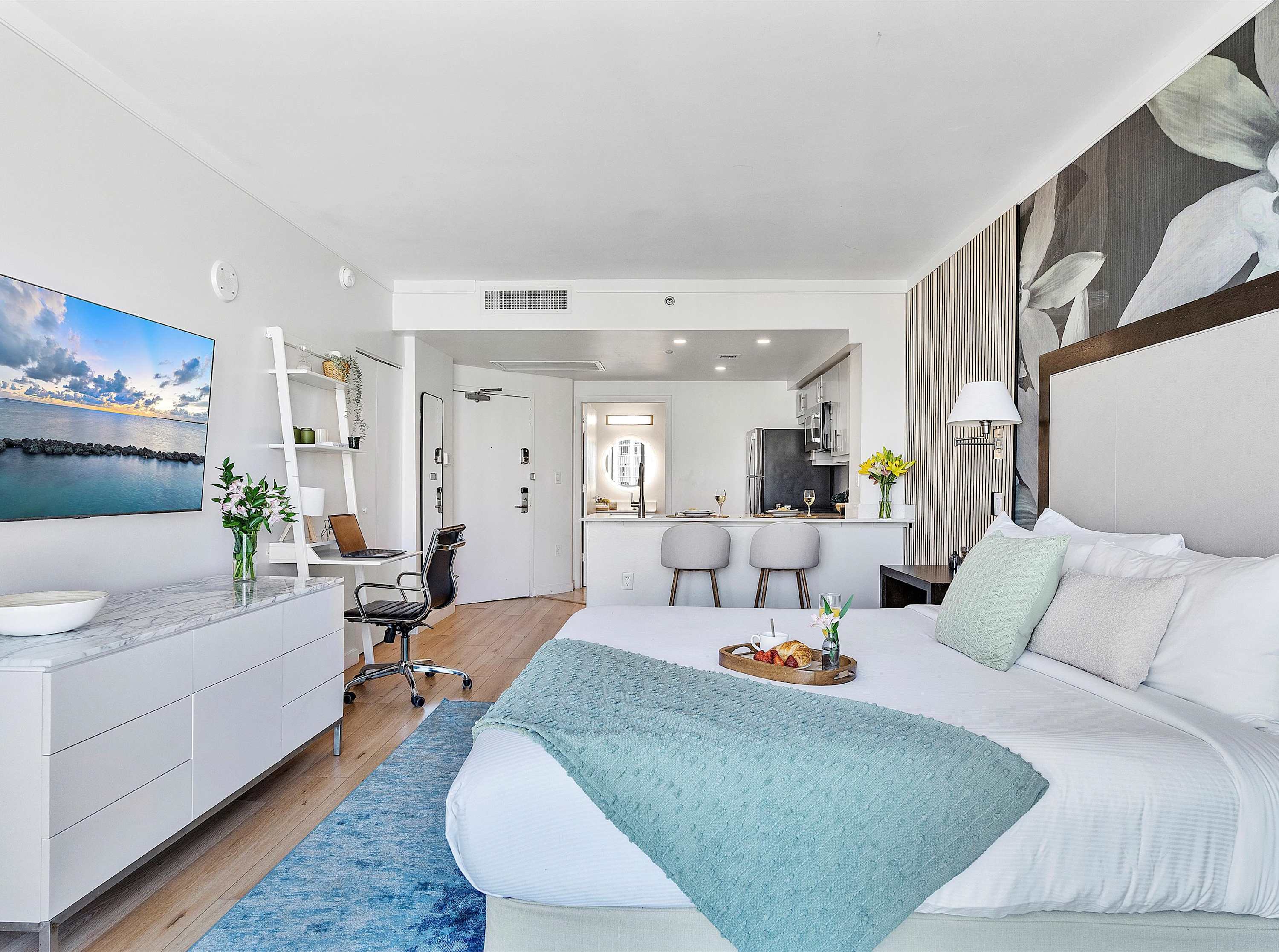A wide view of the studio apartment from the bed, showing the open layout that includes a dedicated workspace, the entrance, and a glimpse of the modern kitchen with a breakfast bar.