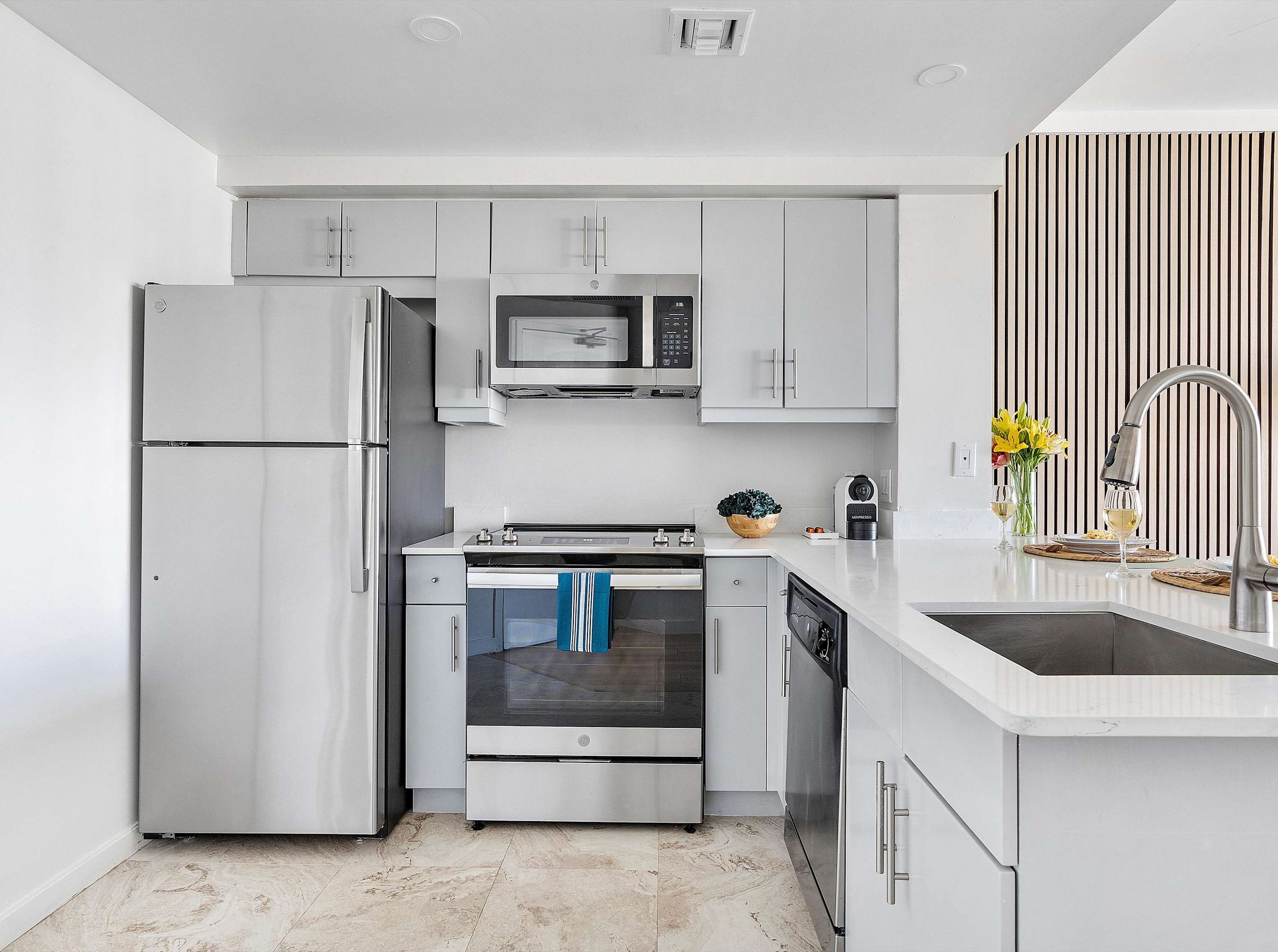 A full view of the contemporary kitchen, equipped with light gray cabinets, stainless steel appliances including a large refrigerator, oven, microwave, and dishwasher, and a white quartz countertop.