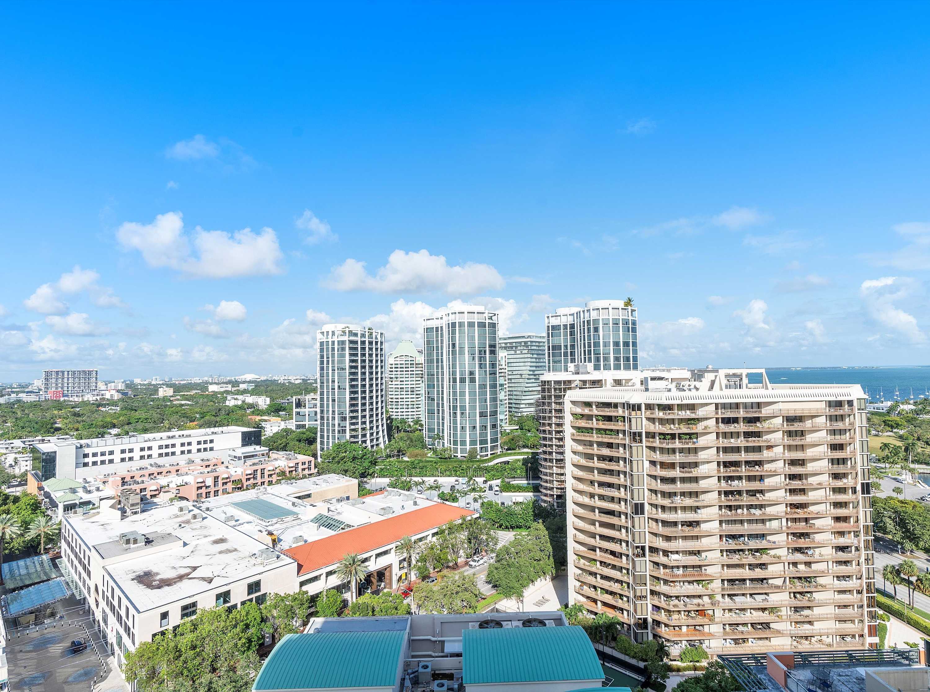 High-angle view of a vibrant city skyline under a clear blue sky with scattered clouds, featuring numerous high-rise buildings, lush green areas, and a glimpse of the ocean in the distance.