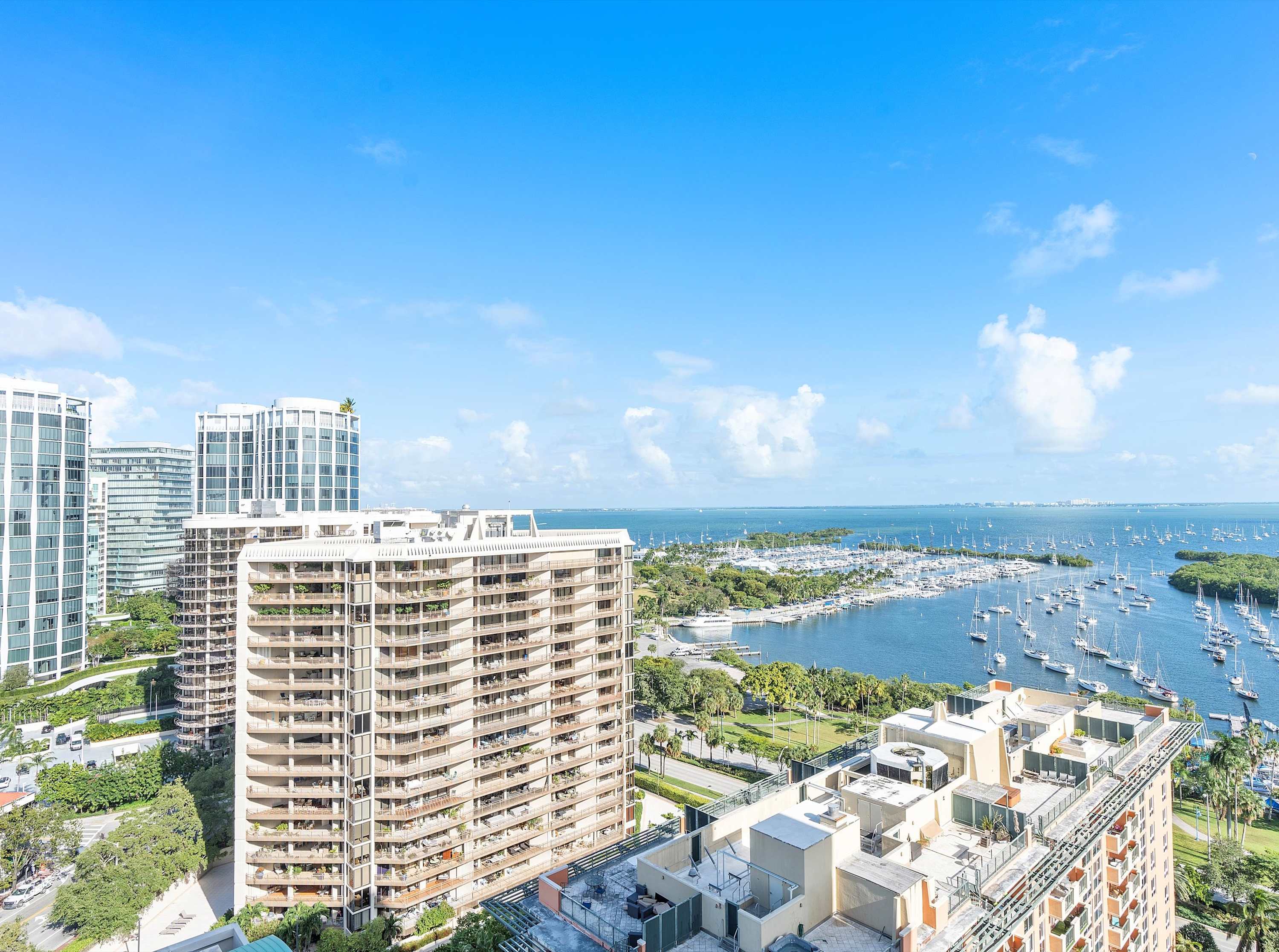 A high-angle exterior view showcasing a vibrant blue bay filled with numerous sailboats and yachts, flanked by lush green trees and several tall residential buildings under a clear sky.