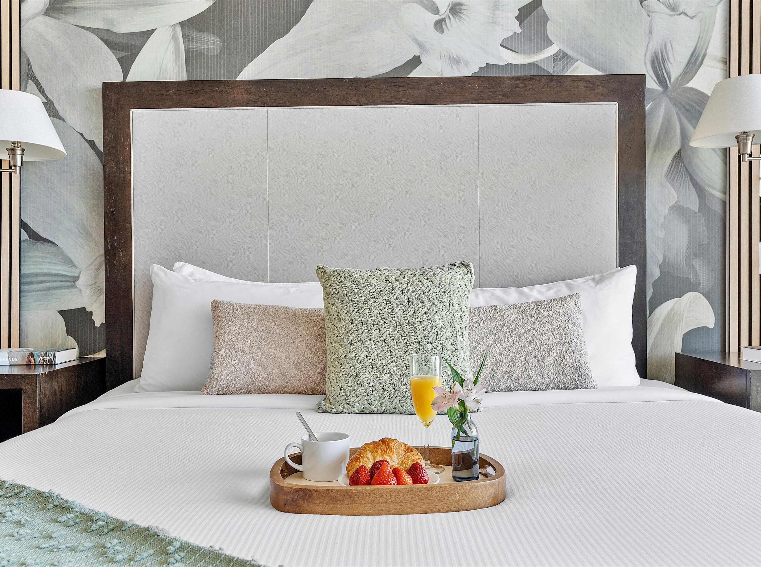 A close-up of the comfortable king-sized bed, adorned with white linens, a light green throw, and a wooden tray holding breakfast items like a croissant, strawberries, and orange juice.
