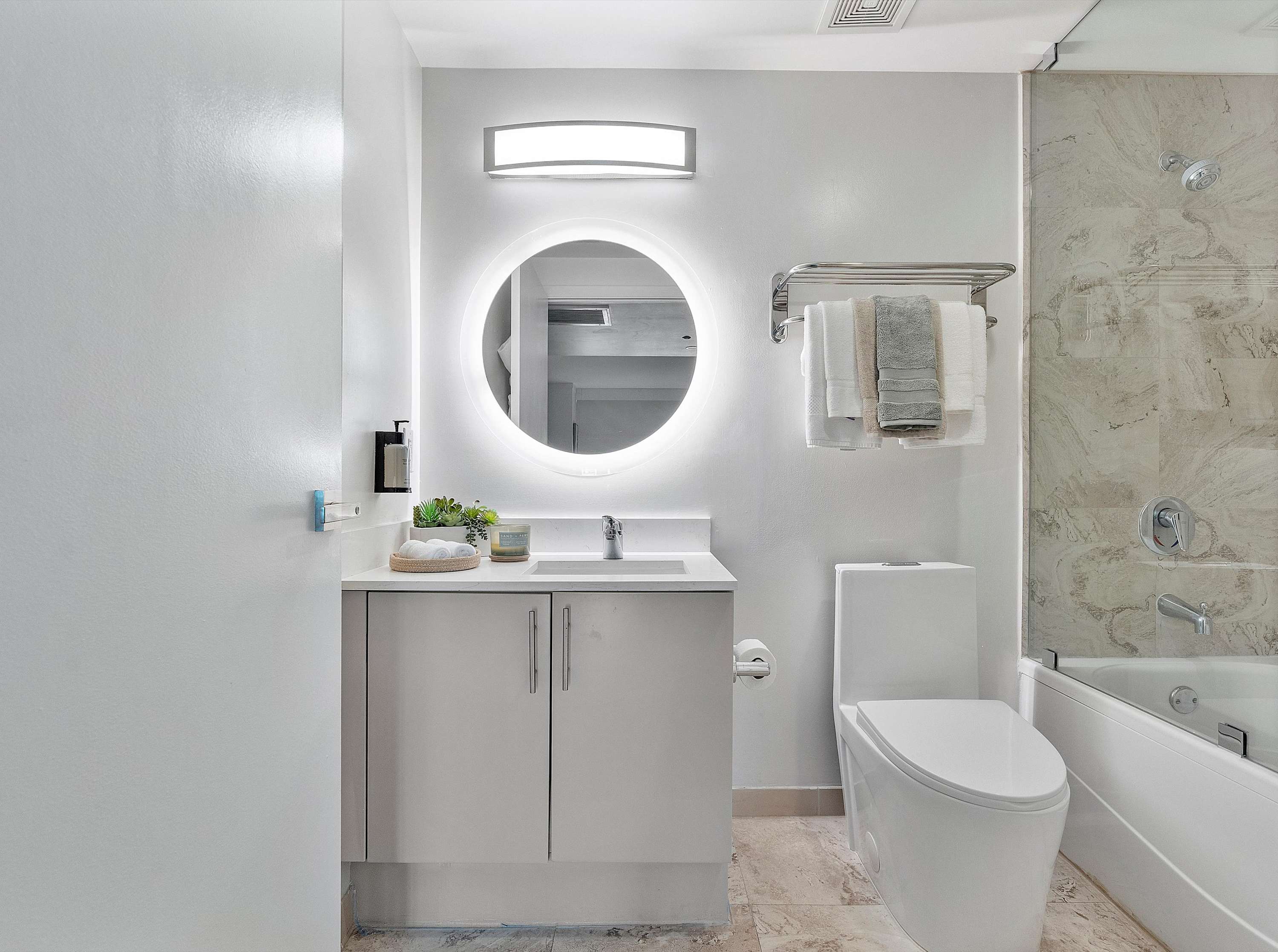 Modern bathroom featuring a white vanity with a light gray cabinet, a round backlit mirror, and a toilet next to a bathtub with a glass shower screen. The walls are light, and the floor is tiled.