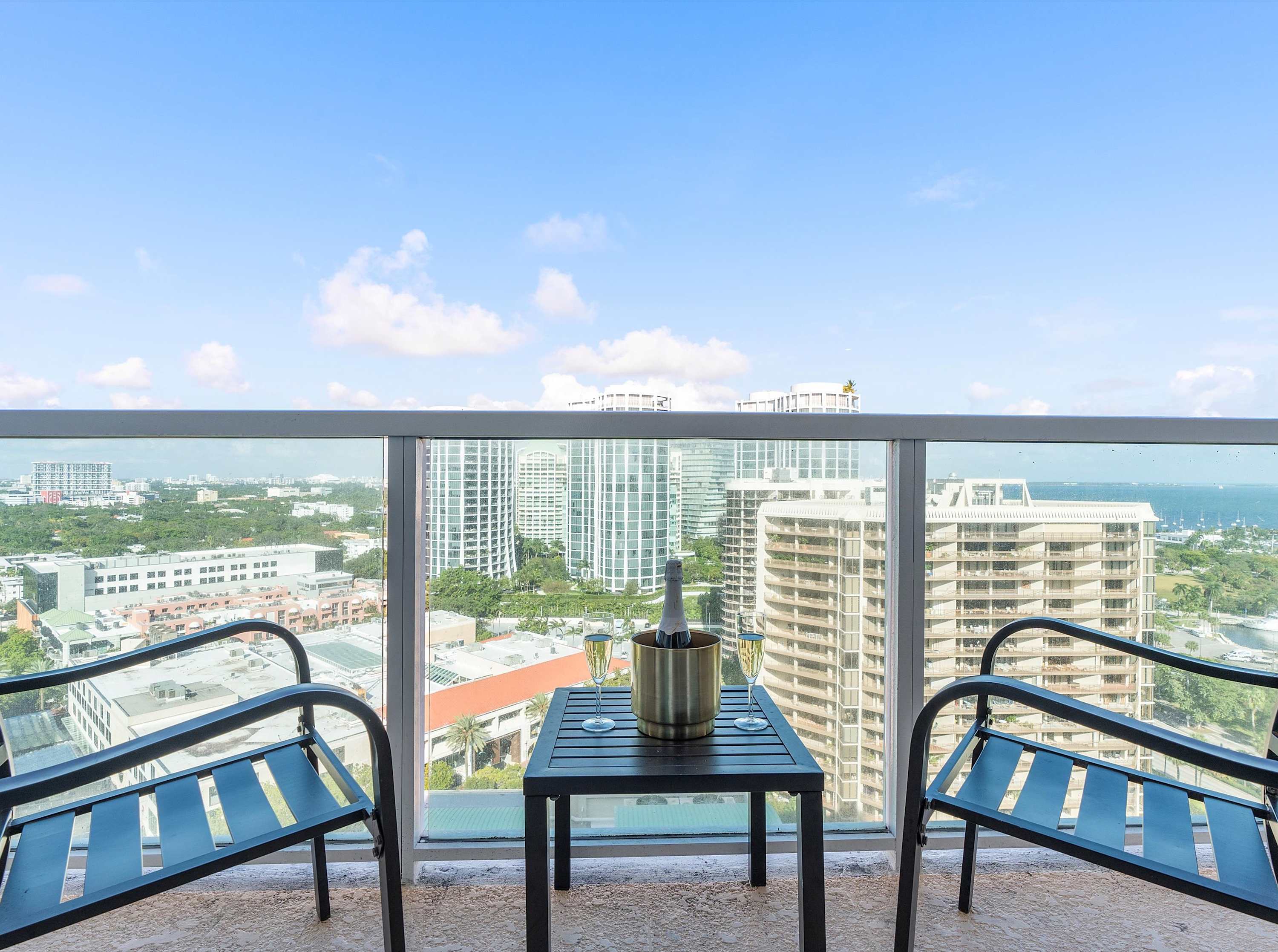 A private balcony furnished with two black chairs and a small table holding a bottle of champagne and two glasses, offering expansive views of surrounding city buildings and a glimpse of the bay.