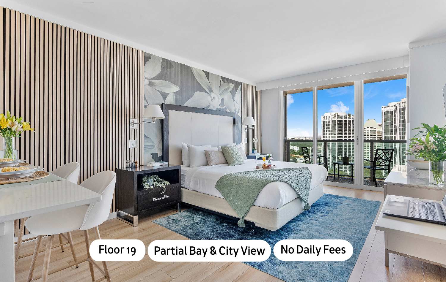 A modern studio apartment featuring a king-sized bed with a floral accent wall, a small dining area, and a private balcony offering partial bay and city views. The room has light wood floors and a blue area rug.