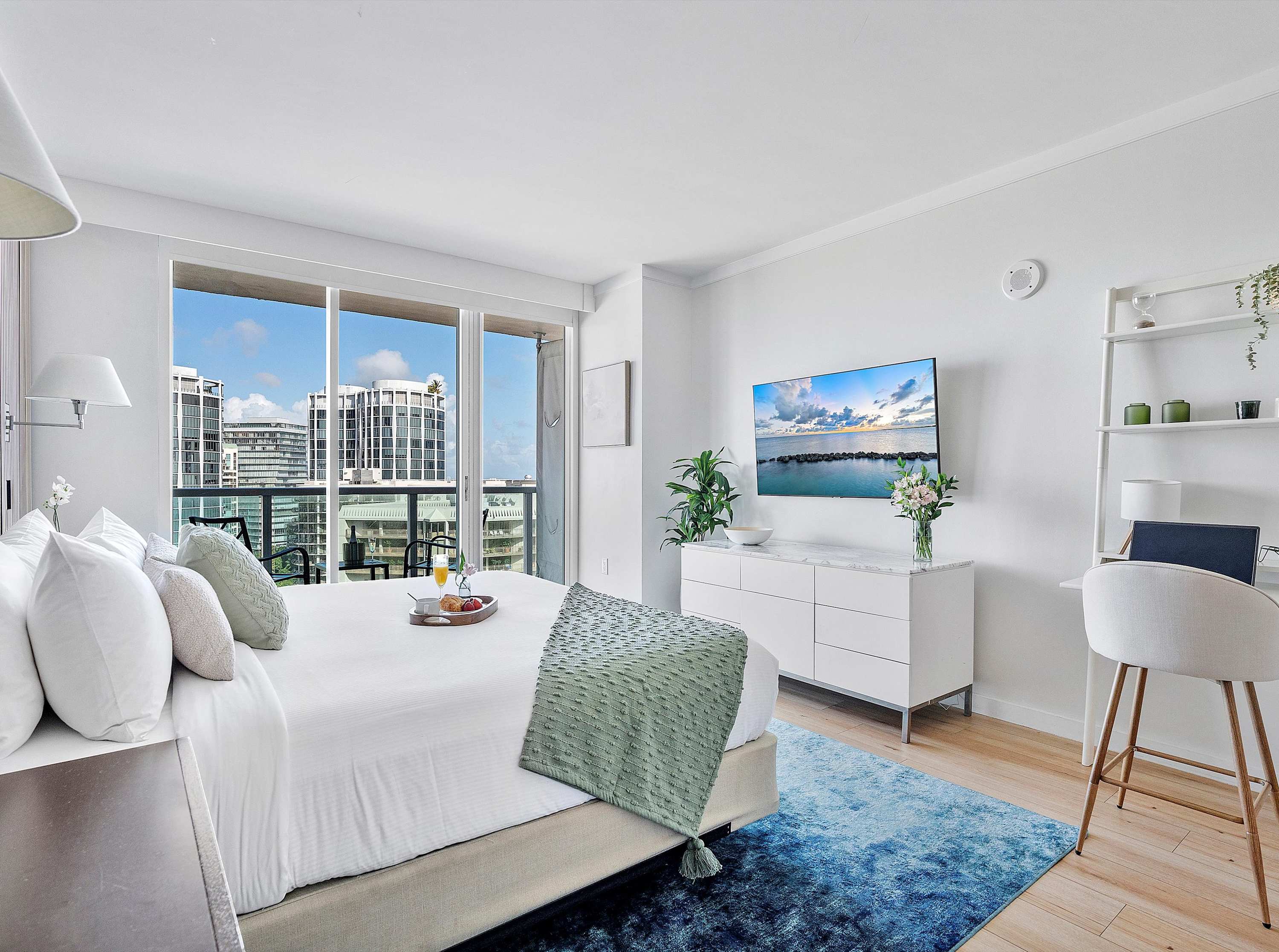 A spacious and bright studio bedroom featuring a king-sized bed, a modern white dresser with a flat-screen TV, a dedicated workspace with a desk, and access to a private balcony with city views.