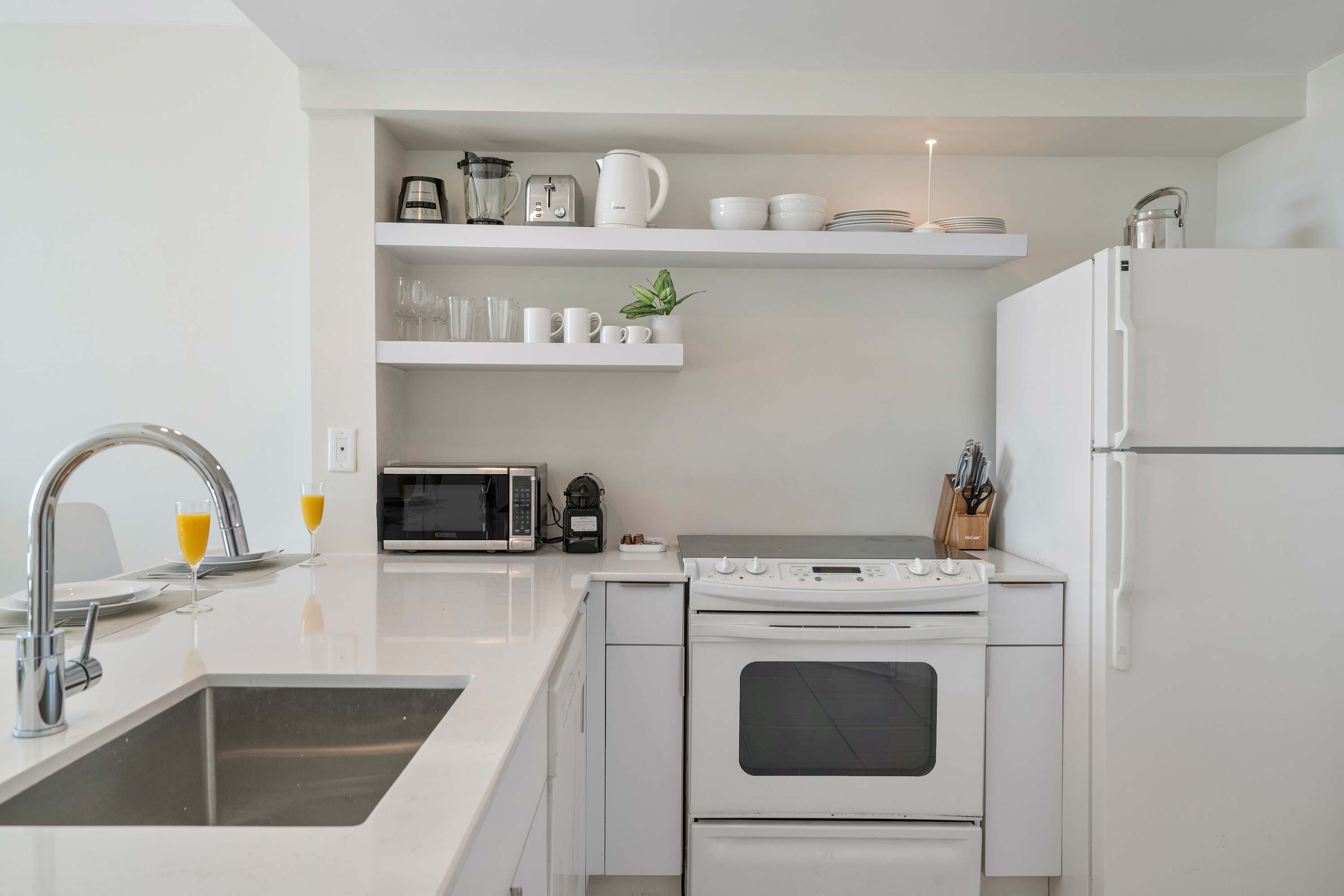Compact, modern kitchen with white cabinetry, white countertops, and open shelving. Appliances include a full-sized refrigerator, electric stove, microwave, toaster, and coffee maker.