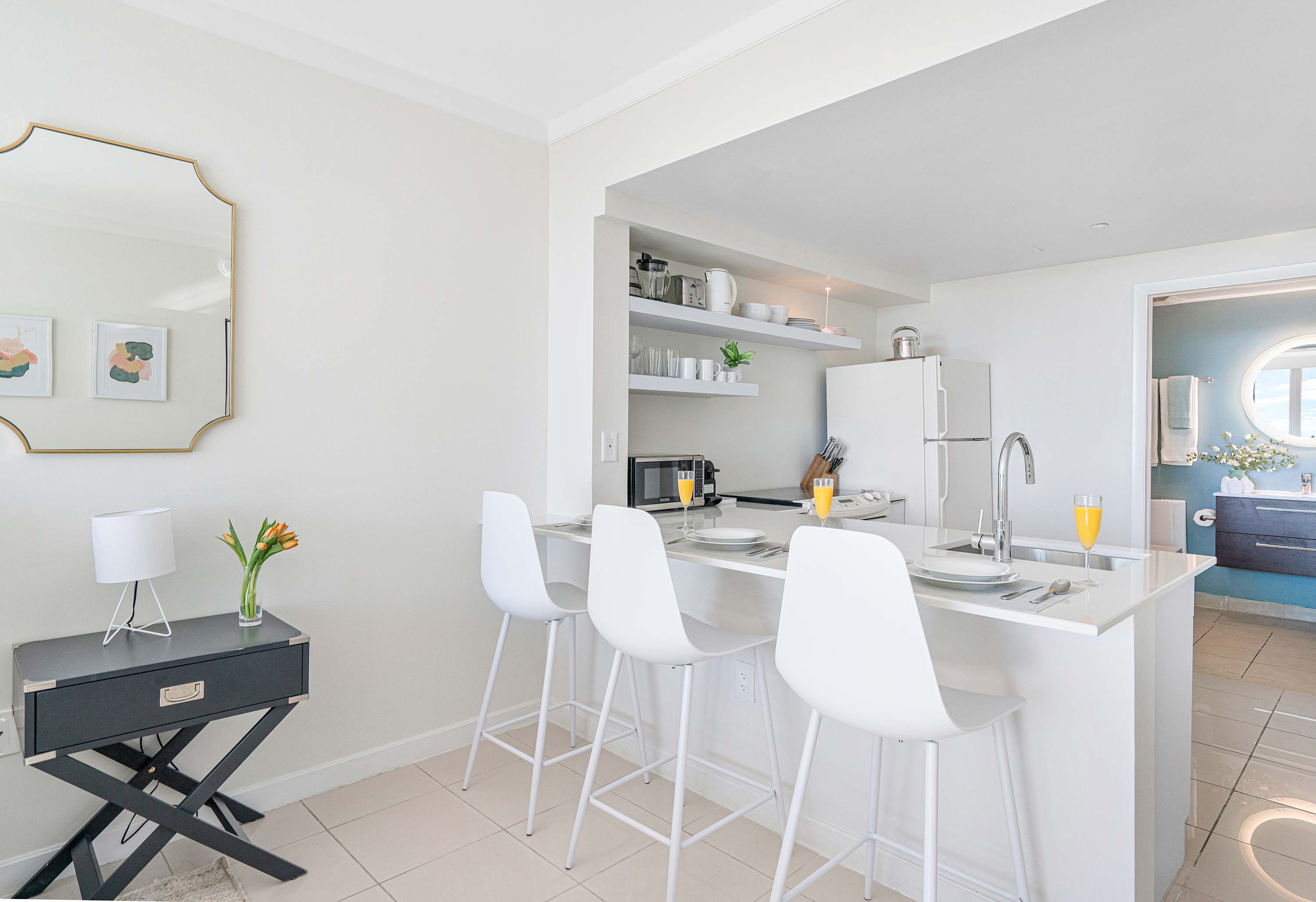 View from the bedroom towards the kitchen and entrance, highlighting the white kitchen island with bar stools. A gold-framed mirror hangs on the wall, and the bathroom entrance is visible in the background.