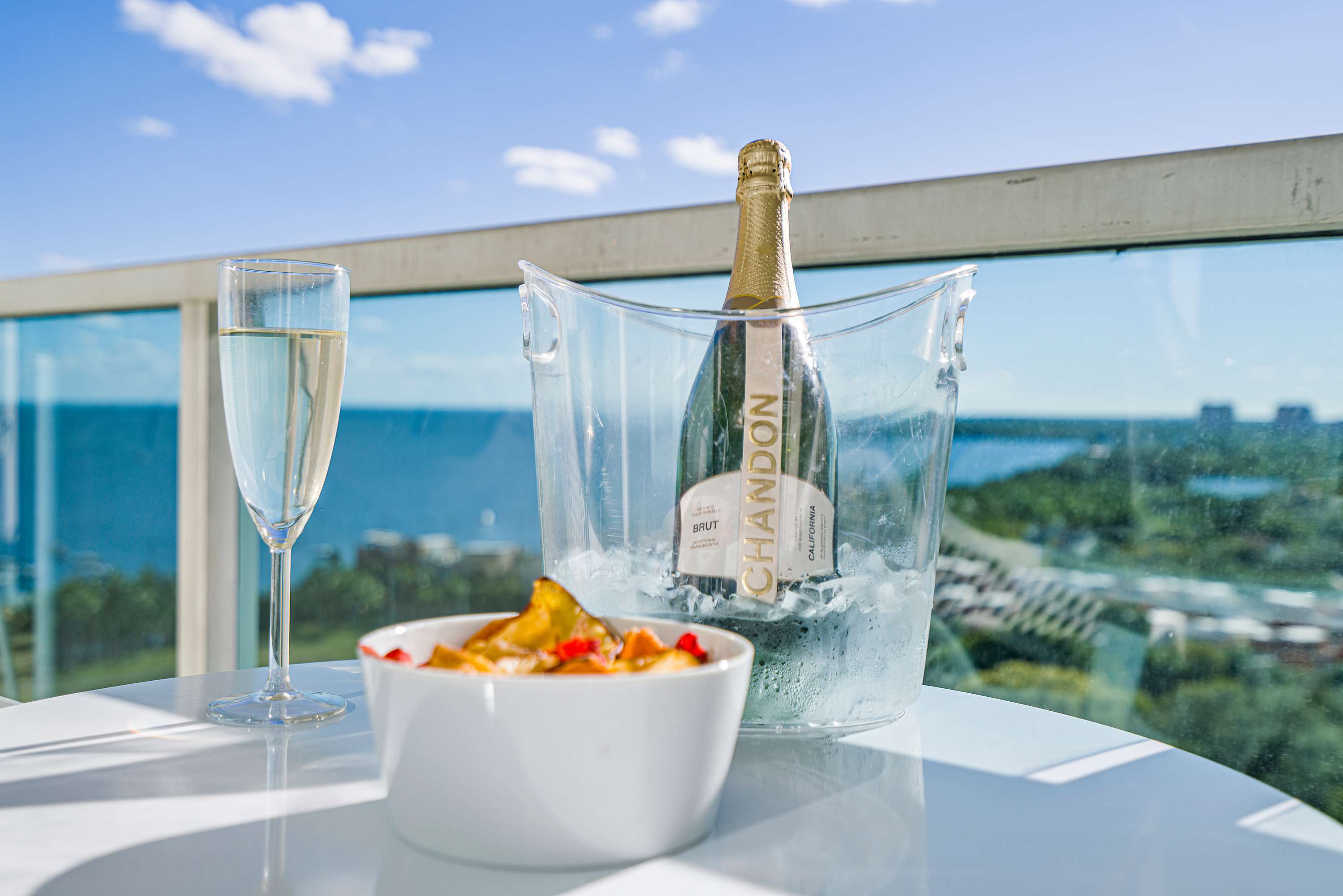 Close-up of a white outdoor table on a balcony, featuring a bottle of chilled champagne in an ice bucket, two champagne flutes, and a bowl of colorful snacks. The sparkling bay is visible in the background.