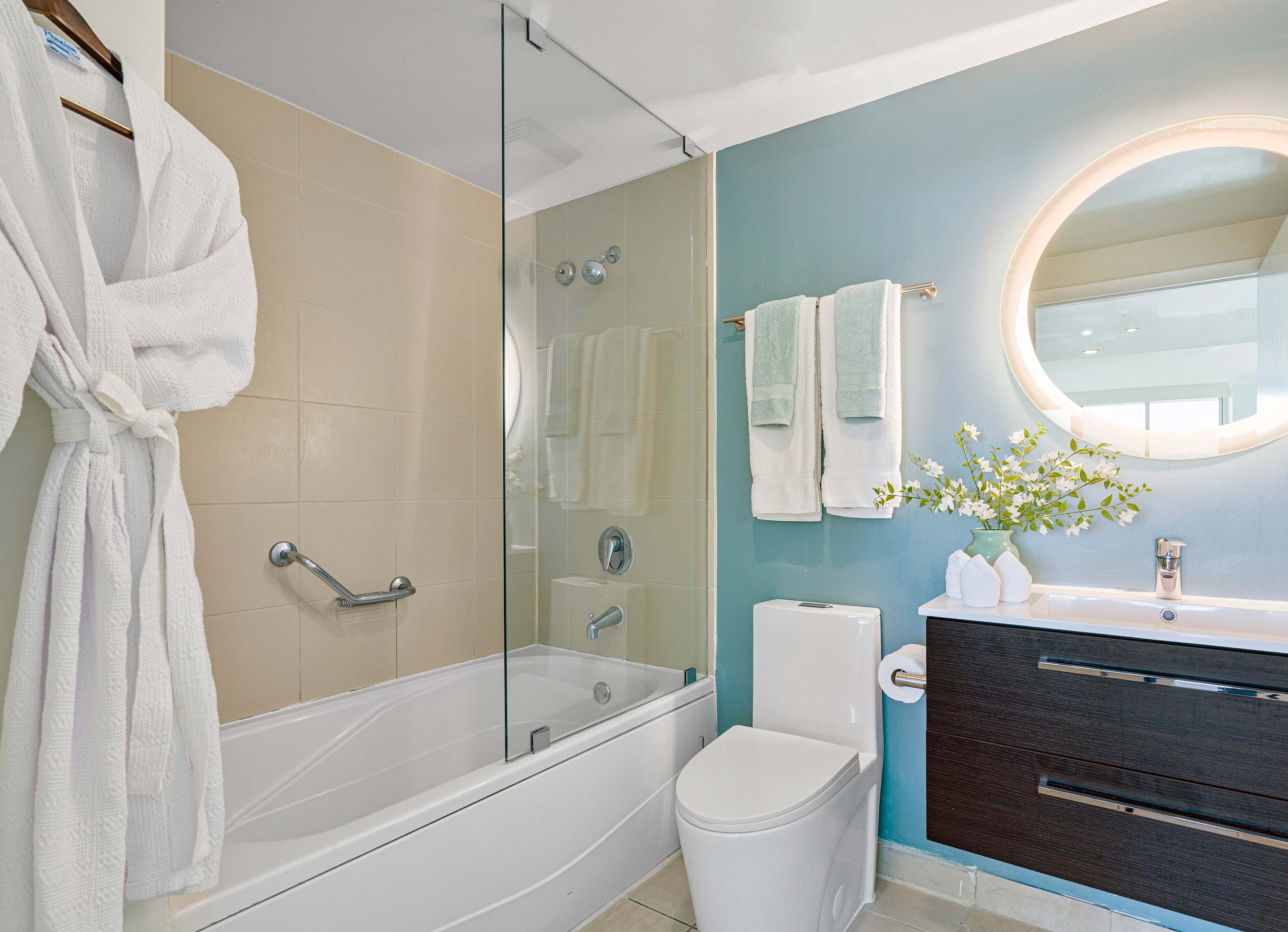 Modern bathroom featuring a bathtub with a glass shower screen, a floating dark wood vanity with a white sink, and a large circular backlit mirror. The walls are tiled in beige and painted light blue, with a white robe hanging on the left.