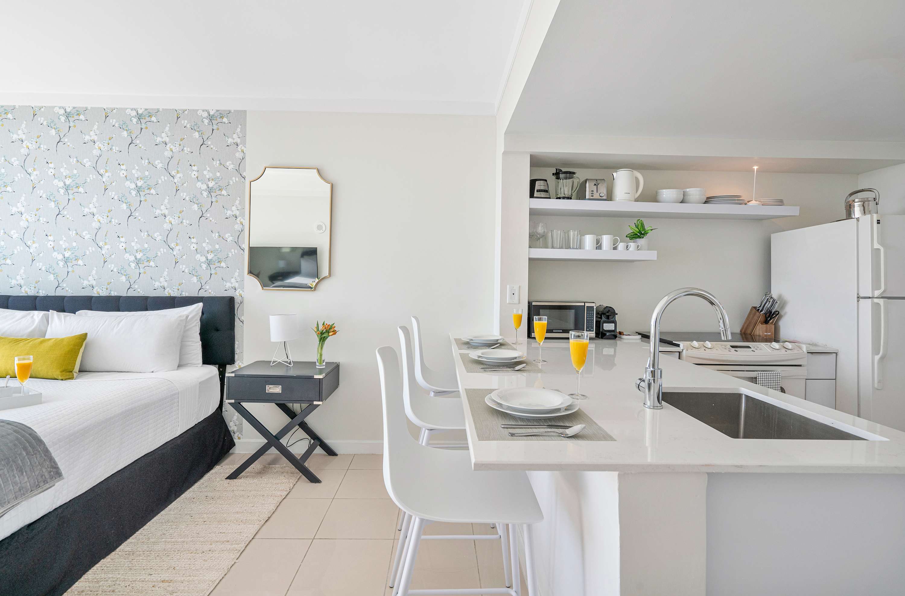 View of the kitchen area from the studio, showing a white kitchen island with three bar stools and a sink. The kitchen features white appliances and open shelving, blending seamlessly with the living space.