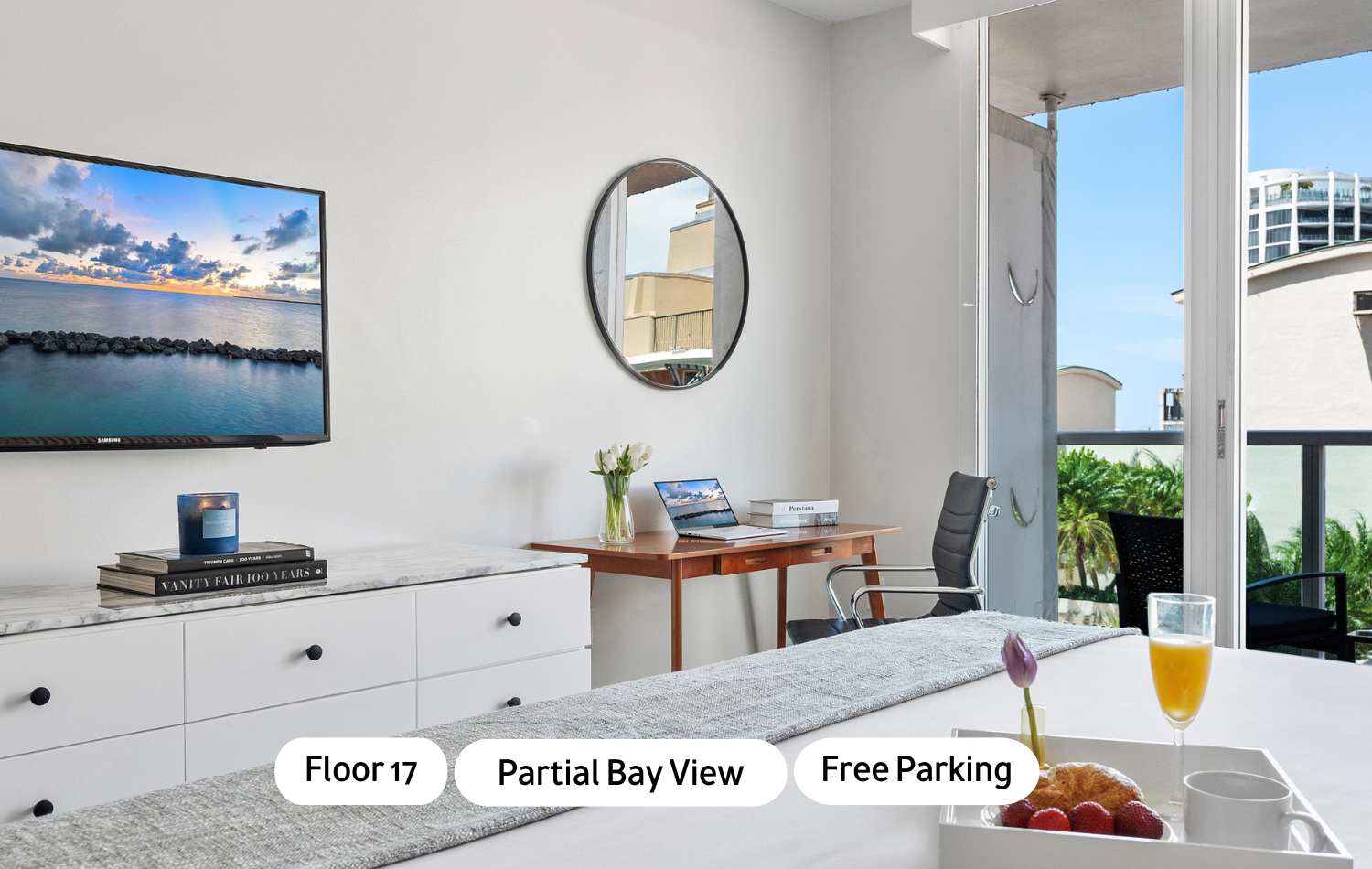 Bright studio room featuring a king bed, white dresser with a flat-screen TV, and a wooden desk with an office chair. A round mirror hangs above the desk, and a balcony with a partial bay view is visible through the glass door.