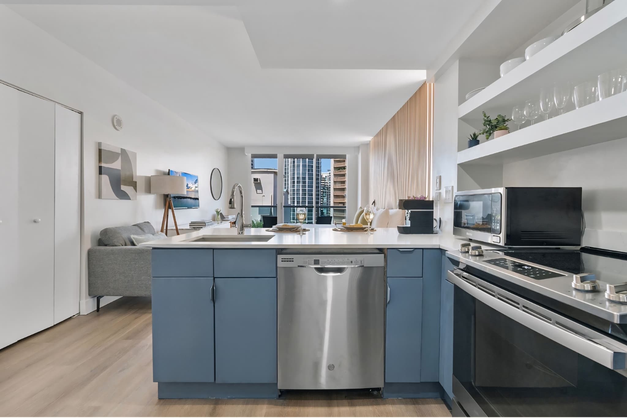 Modern kitchen area featuring light blue lower cabinets, white quartz countertops, a stainless steel dishwasher, and a large sink with a gooseneck faucet. The open-concept layout connects to the main living space.