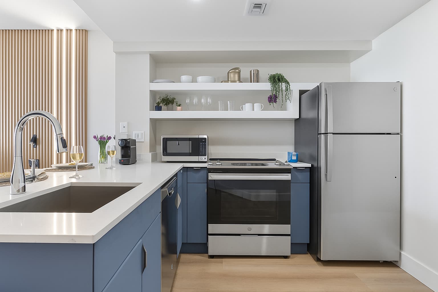 Well-equipped modern kitchen with light blue base cabinets, white countertops, and stainless steel appliances including a refrigerator, oven, and microwave. White open shelving provides additional storage and display space.