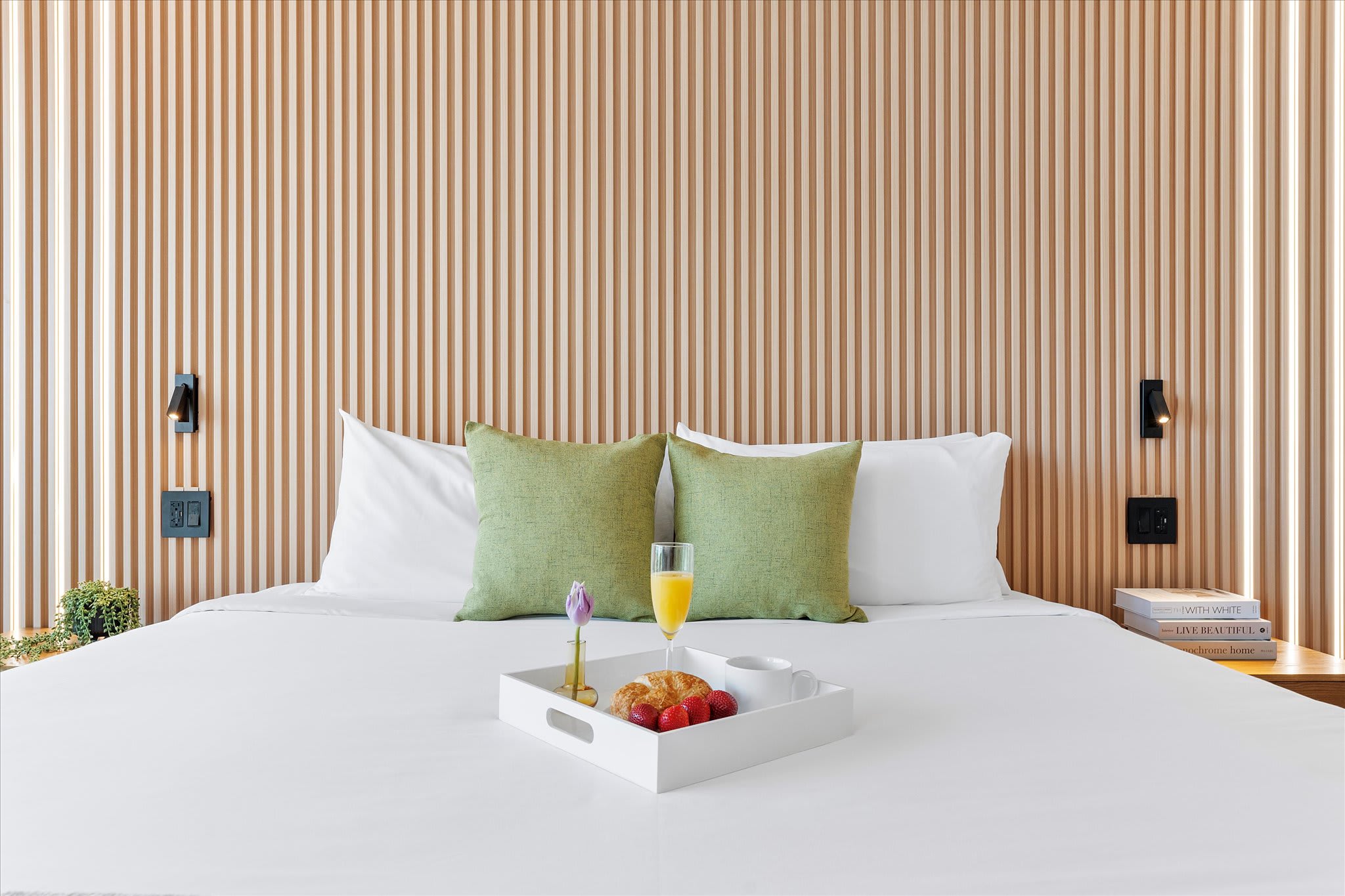 Close-up of a king bed with crisp white linens and two green throw pillows, set against a light wood slatted accent wall with integrated vertical lighting. A white tray with breakfast items rests on the bed.