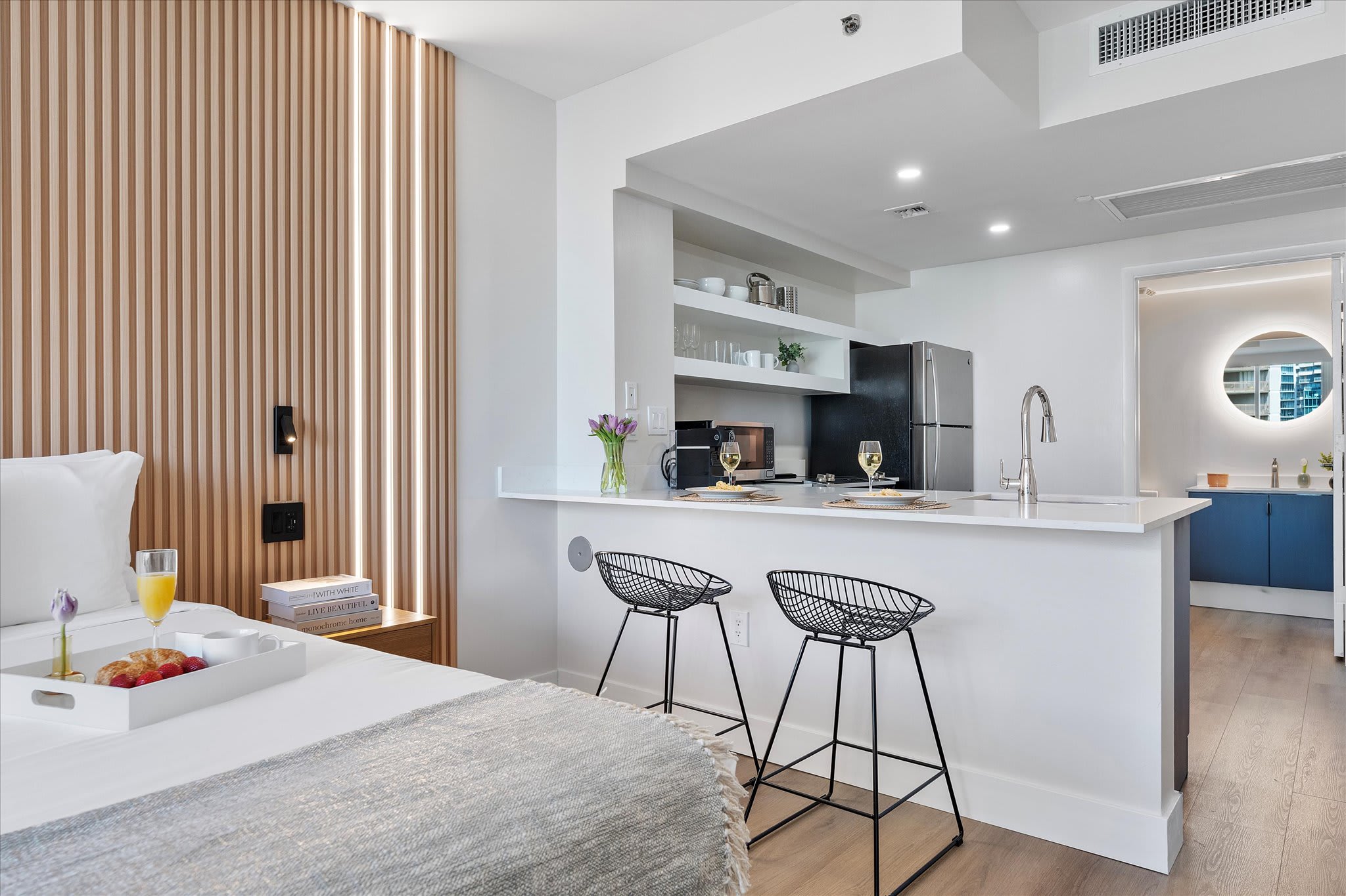 Modern kitchen with light blue lower cabinets, white countertops, stainless steel appliances, and open shelving. A breakfast bar with two black wire bar stools separates the kitchen from the sleeping area, which features a slatted wooden accent wall.
