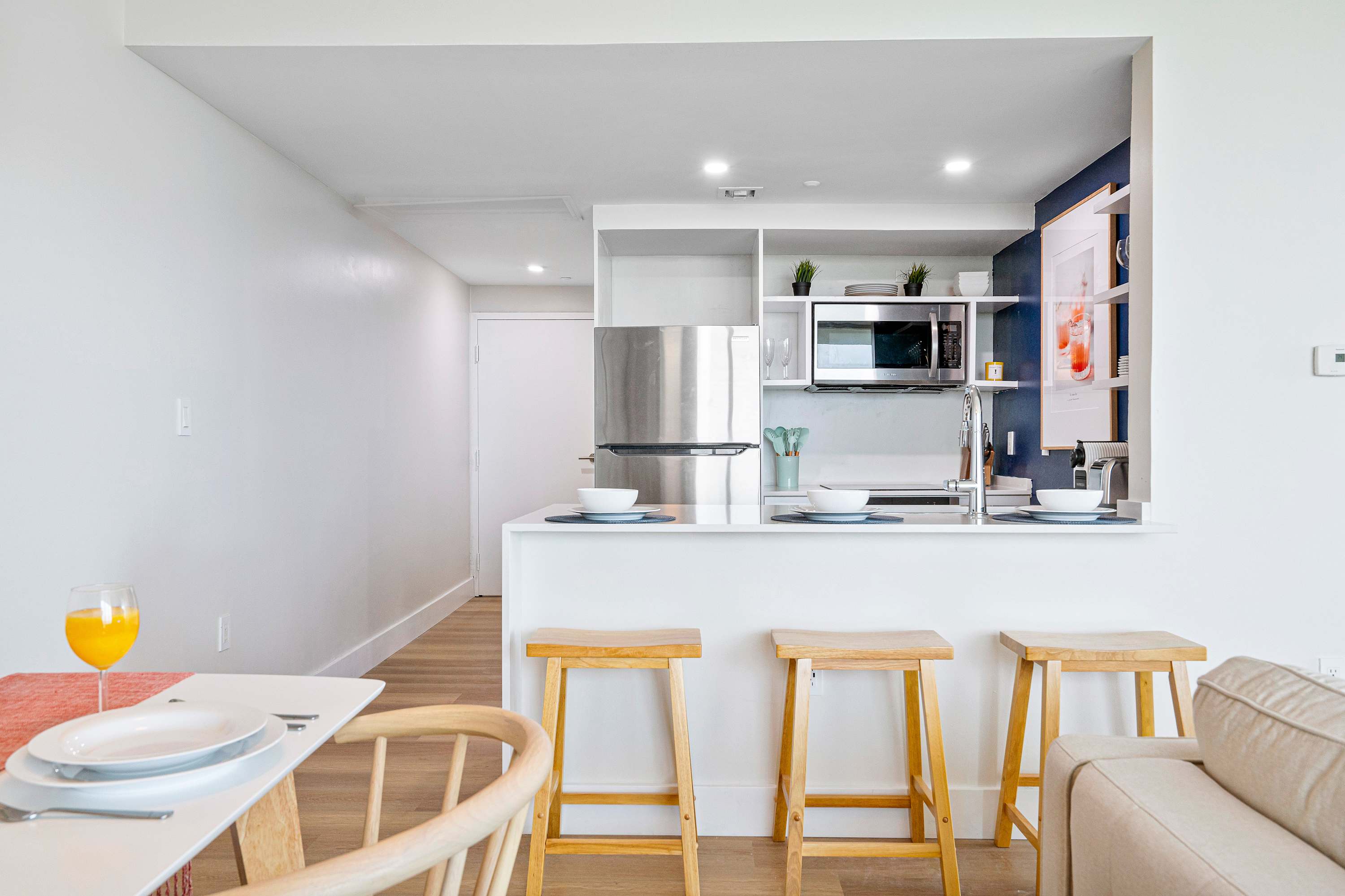 Modern kitchen with a white countertop island, three wooden bar stools, and stainless steel appliances including a refrigerator and microwave, seen from the dining area.
