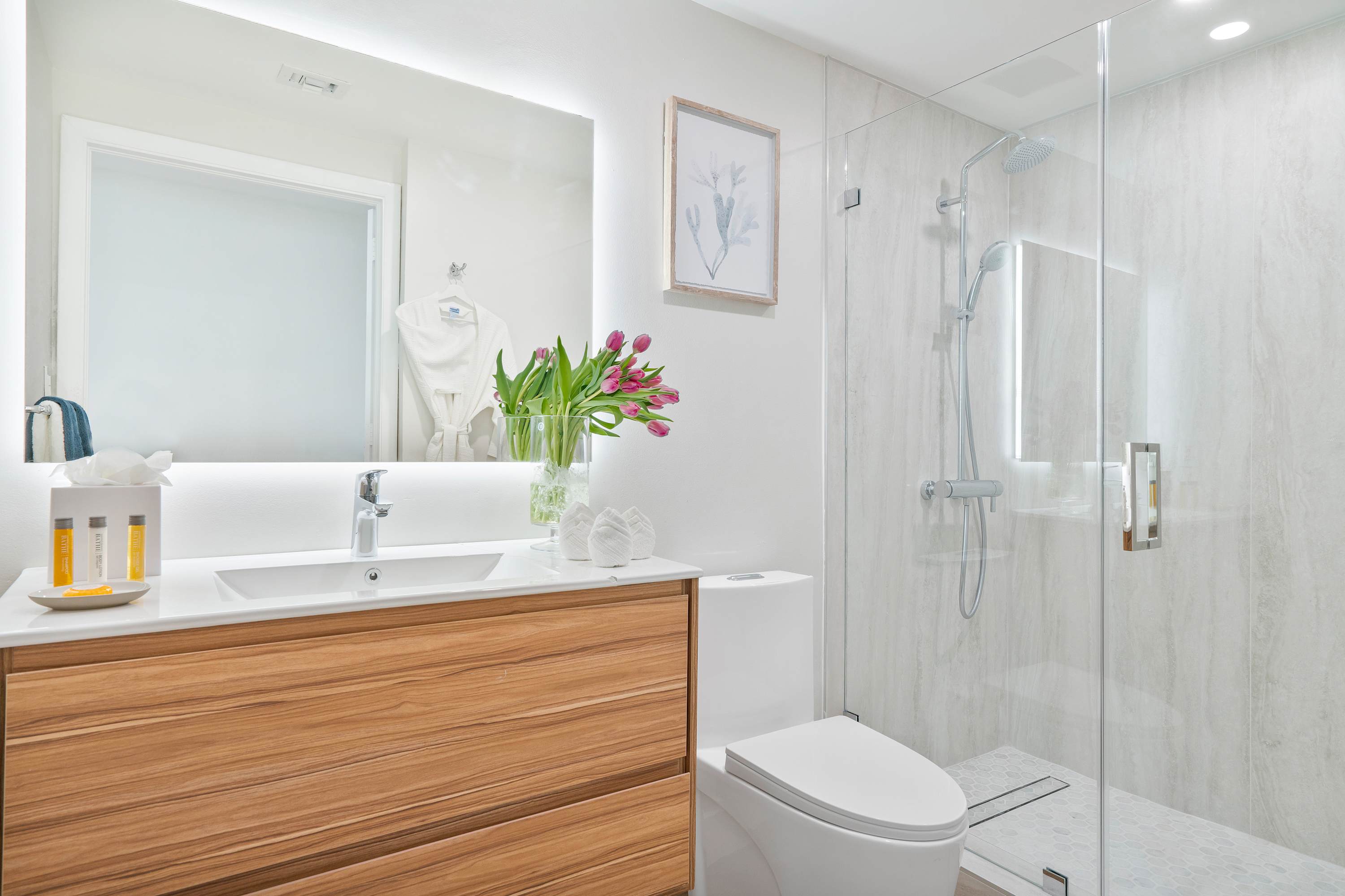 Modern bathroom featuring a wooden vanity with a white countertop and rectangular sink, adjacent to a walk-in glass shower with light-colored tiles and a toilet.
