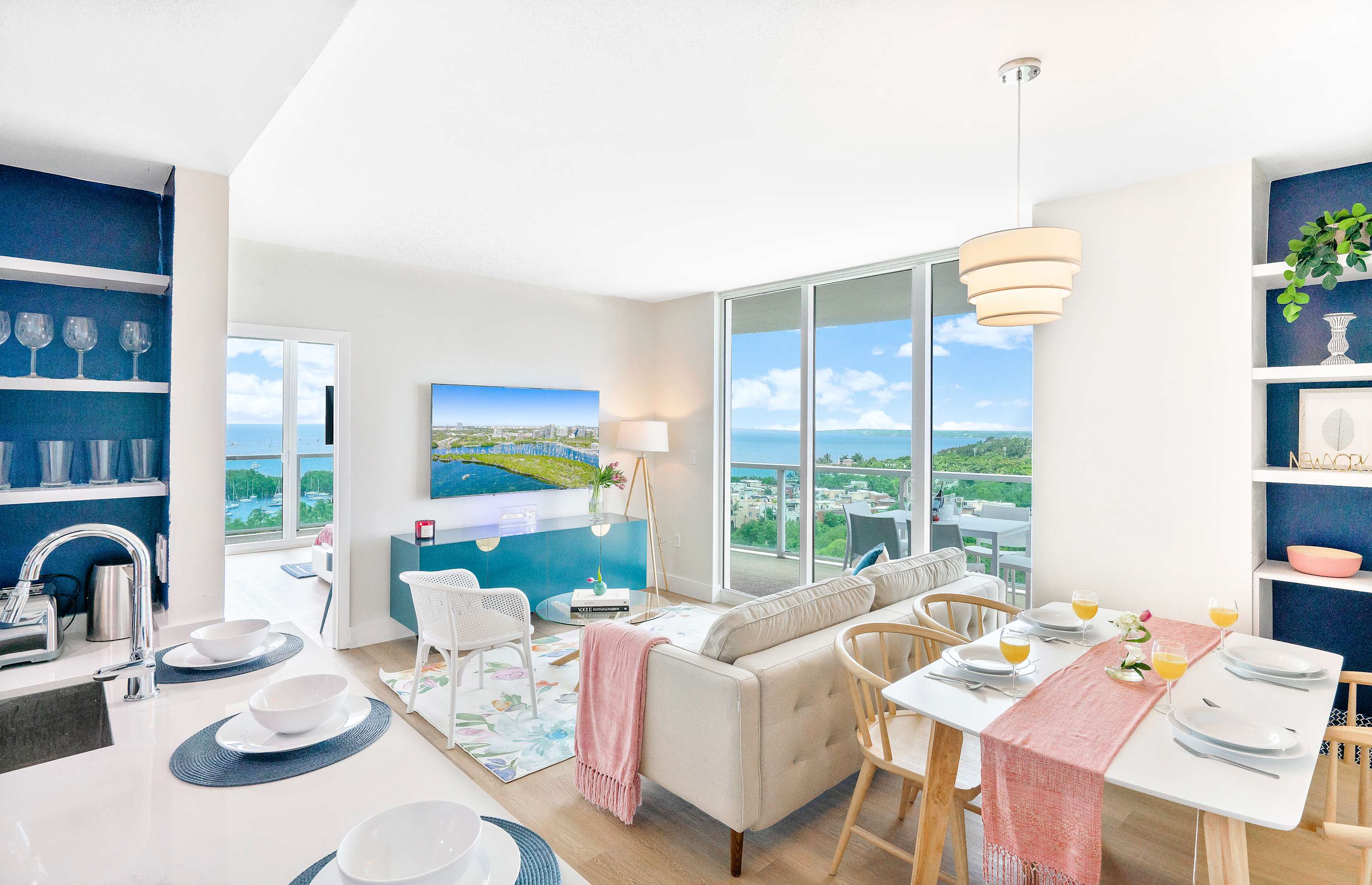 Open-concept living and dining area with a light-colored sofa, a dining table set for four, and large sliding glass doors leading to a balcony with a panoramic sea view.