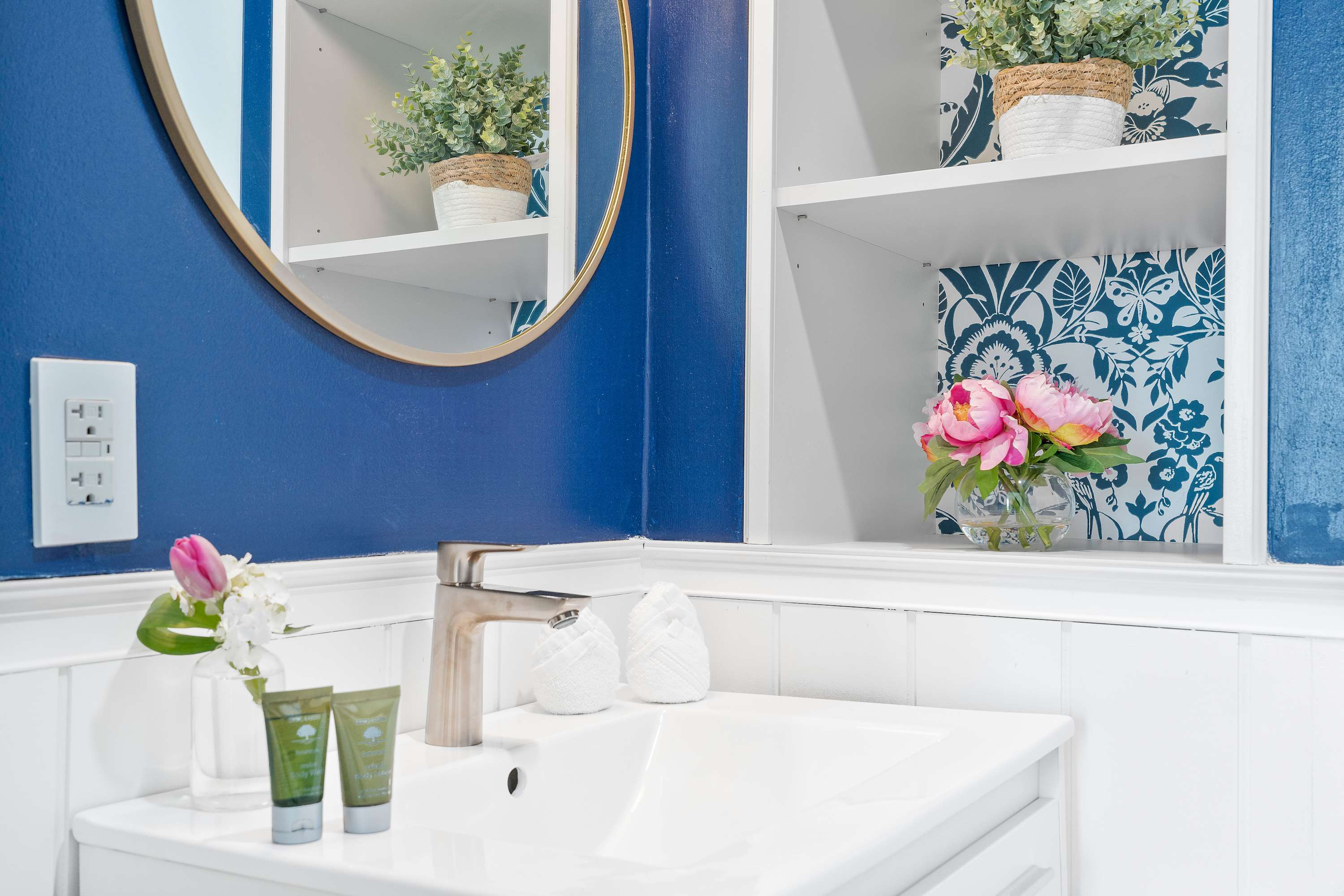 Modern bathroom with a white vanity, a vessel sink, and a round gold-framed mirror, featuring a blue accent wall and white wainscoting.