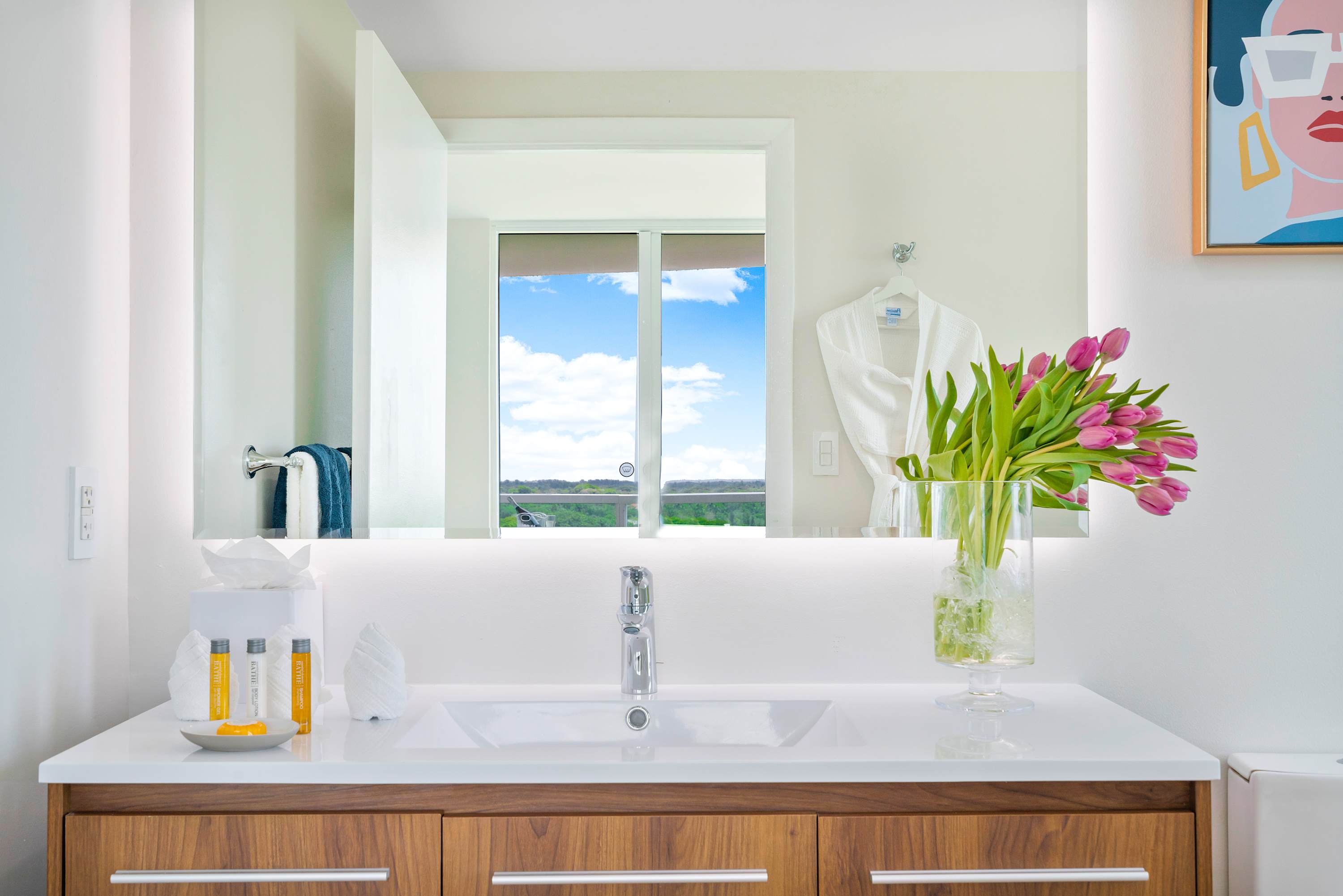 Modern bathroom vanity with a white countertop, wooden drawers, a rectangular sink, and a large mirror reflecting a window with a bright sky and distant view. Pink tulips and toiletries adorn the counter.
