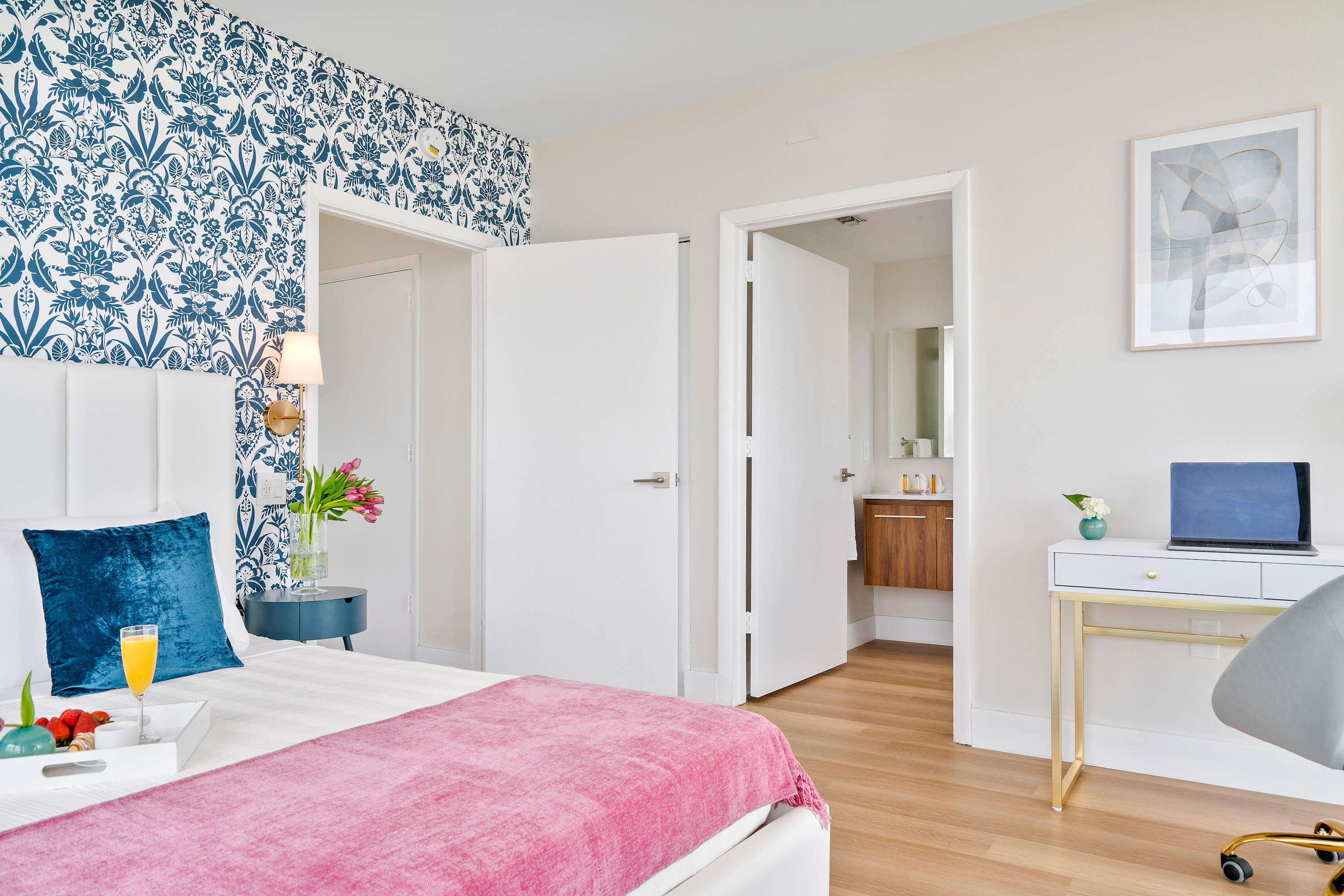 Bright bedroom with a white upholstered bed, pink throw blanket, and blue patterned accent wall. A small desk with a laptop is visible, and an open door leads to a bathroom vanity.