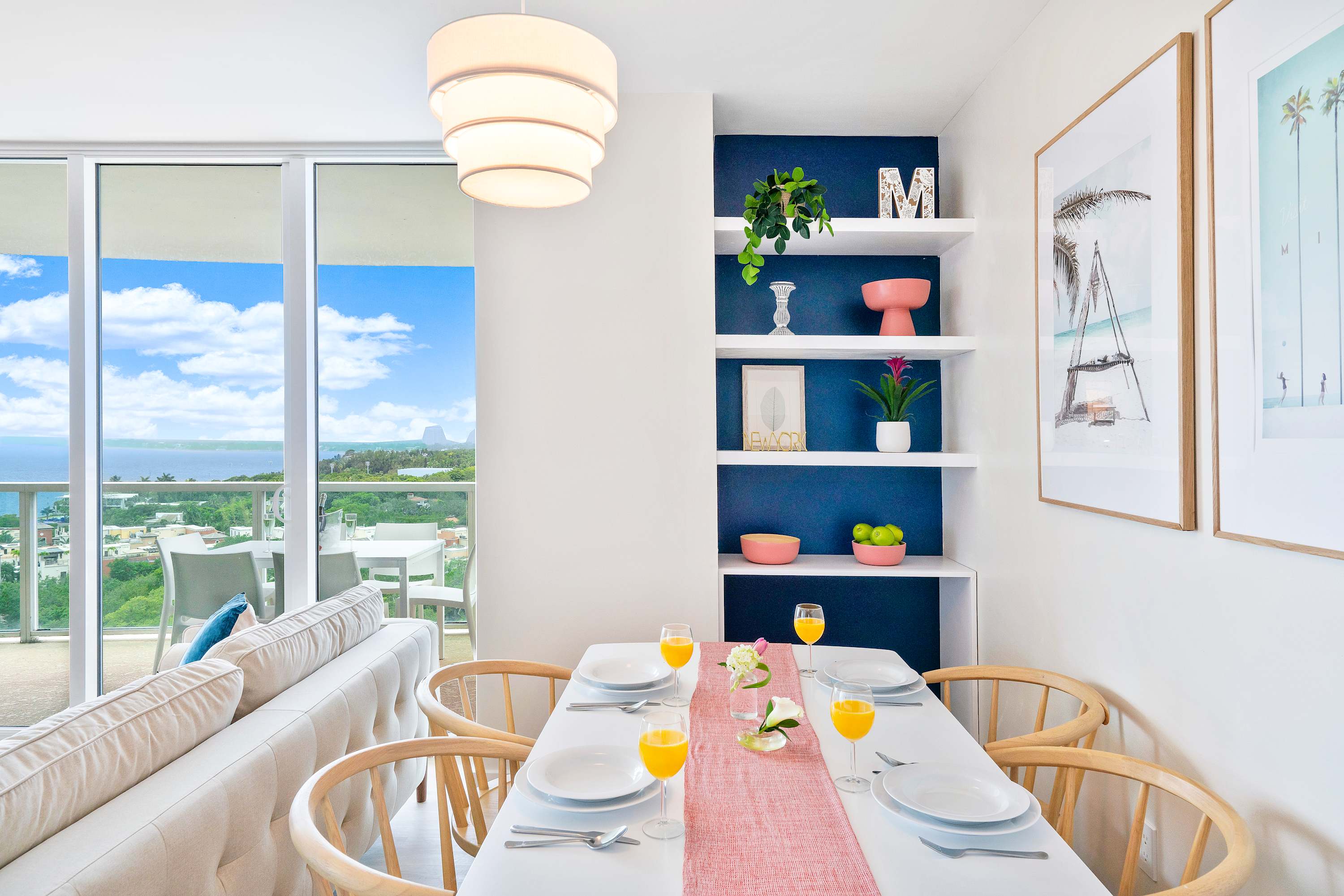 Bright dining area with a white table, four wooden chairs, and a pink runner, featuring a built-in navy blue shelving unit with decorative items. Large sliding glass doors open to a balcony with expansive ocean and city views.