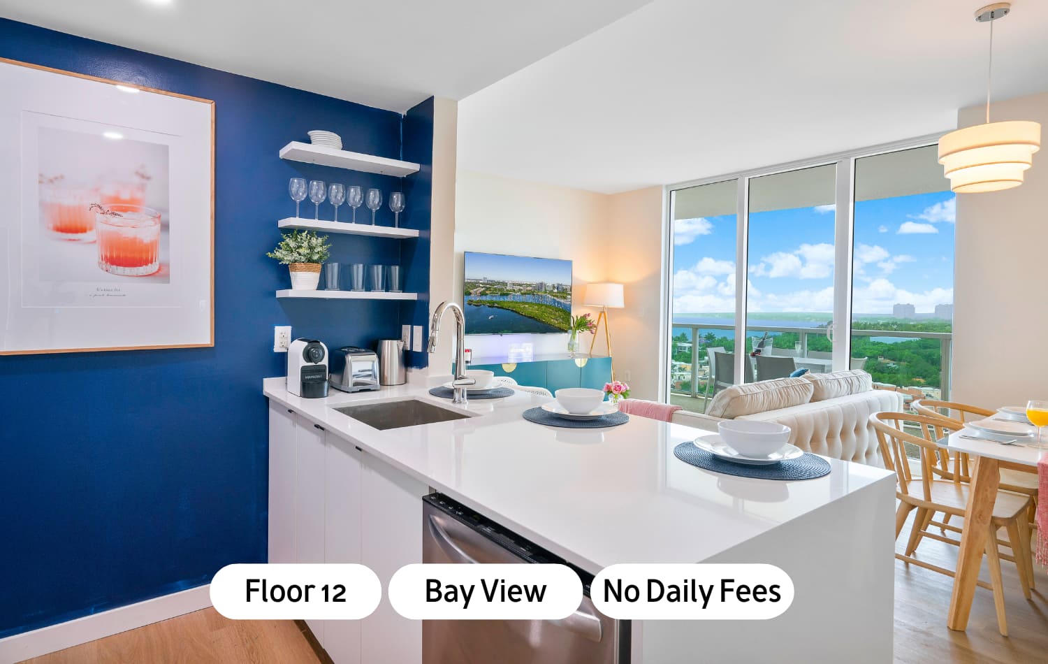 Modern kitchen with a white quartz island and stainless steel appliances, featuring a bold blue accent wall with open shelving and a view of the bay from the adjacent living area.