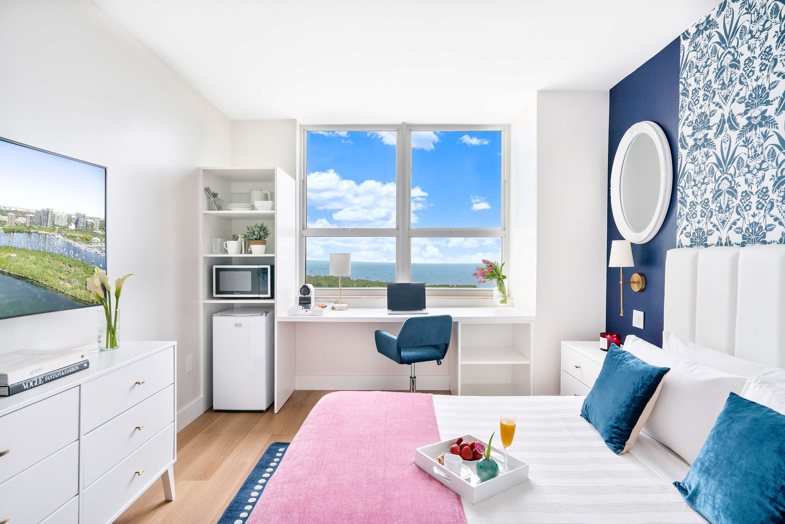 Wide shot of the bedroom showcasing the white king bed with pink throw, a blue and white patterned accent wall, and a desk area with expansive ocean views.