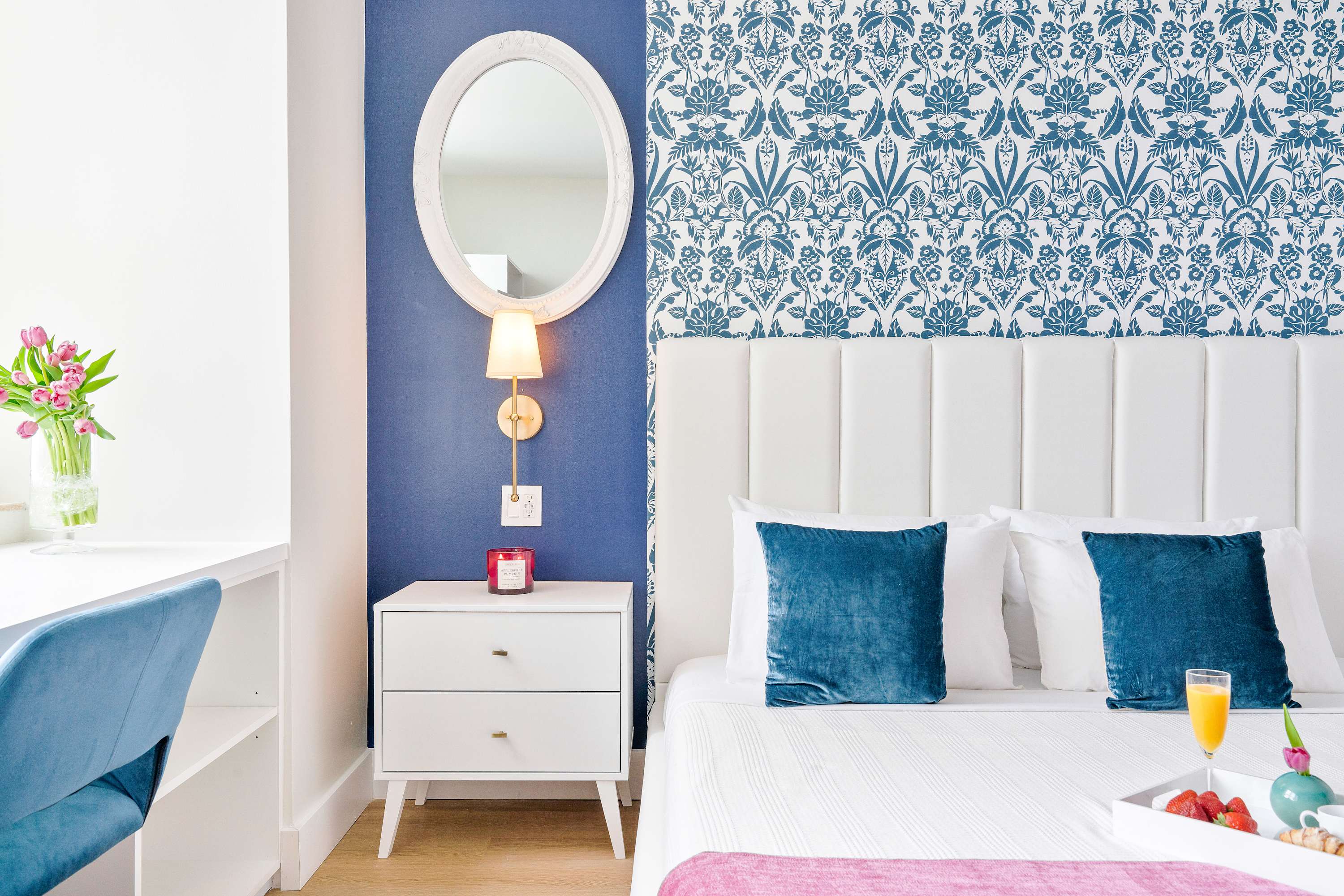 Close-up of the bedroom's white upholstered bed and nightstand, featuring a blue accent wall with a patterned section, an oval mirror, and a wall sconce.