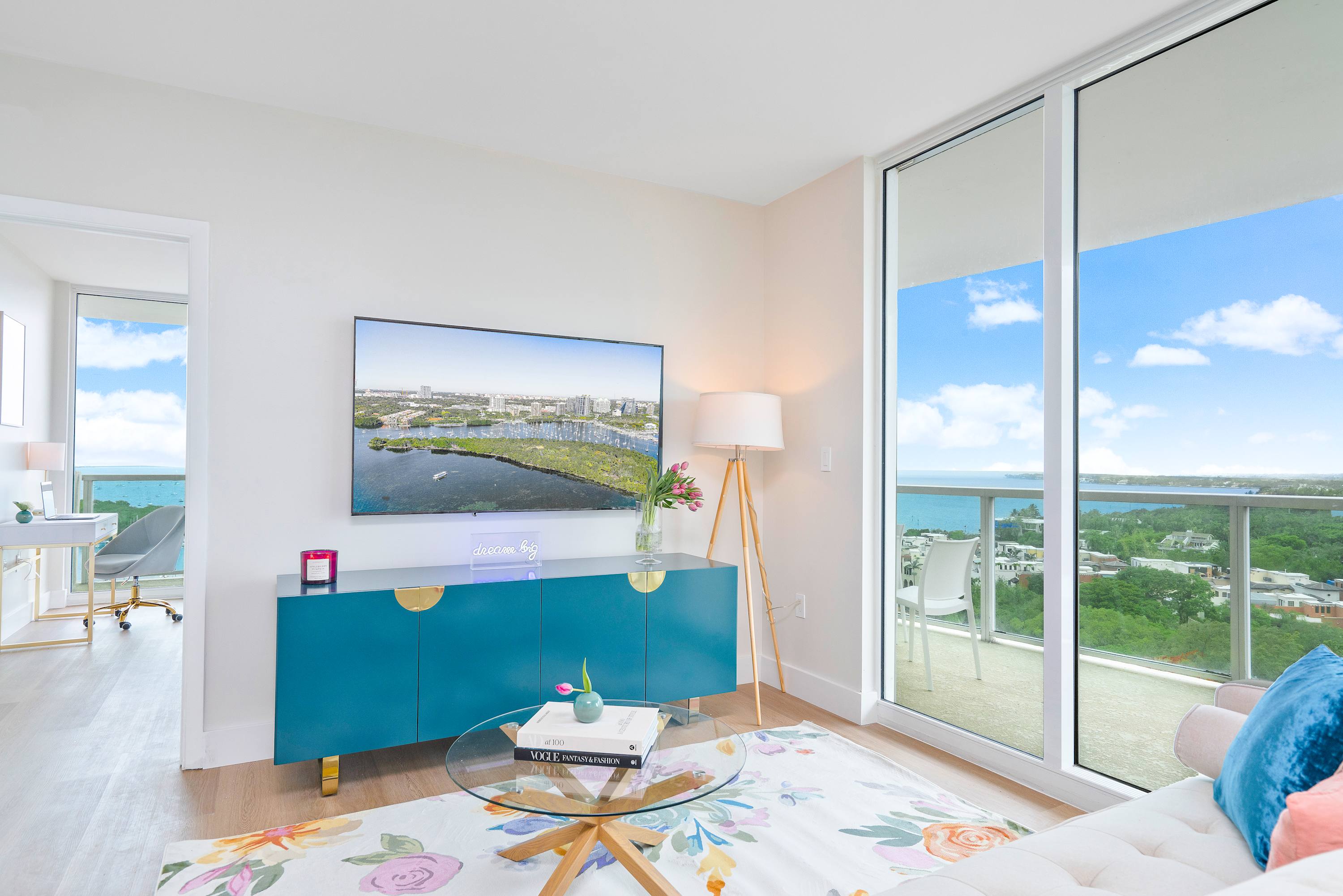 Bright living room with a large flat-screen TV on a teal console, a round glass coffee table, and floor-to-ceiling windows offering expansive ocean views.