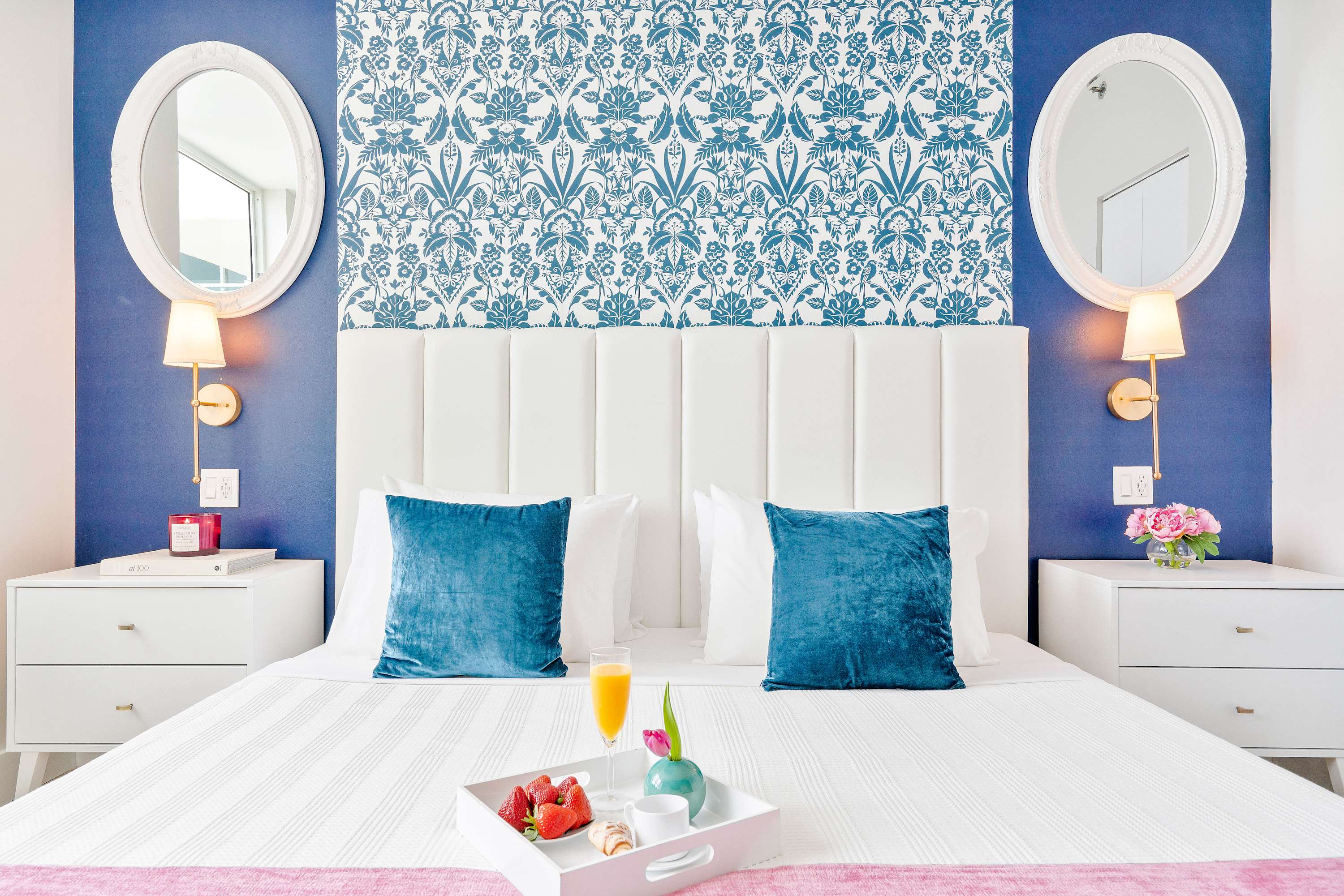 Stylish bedroom with a white upholstered headboard, a bold blue accent wall, and a patterned wallpaper section, flanked by white nightstands and oval mirrors.