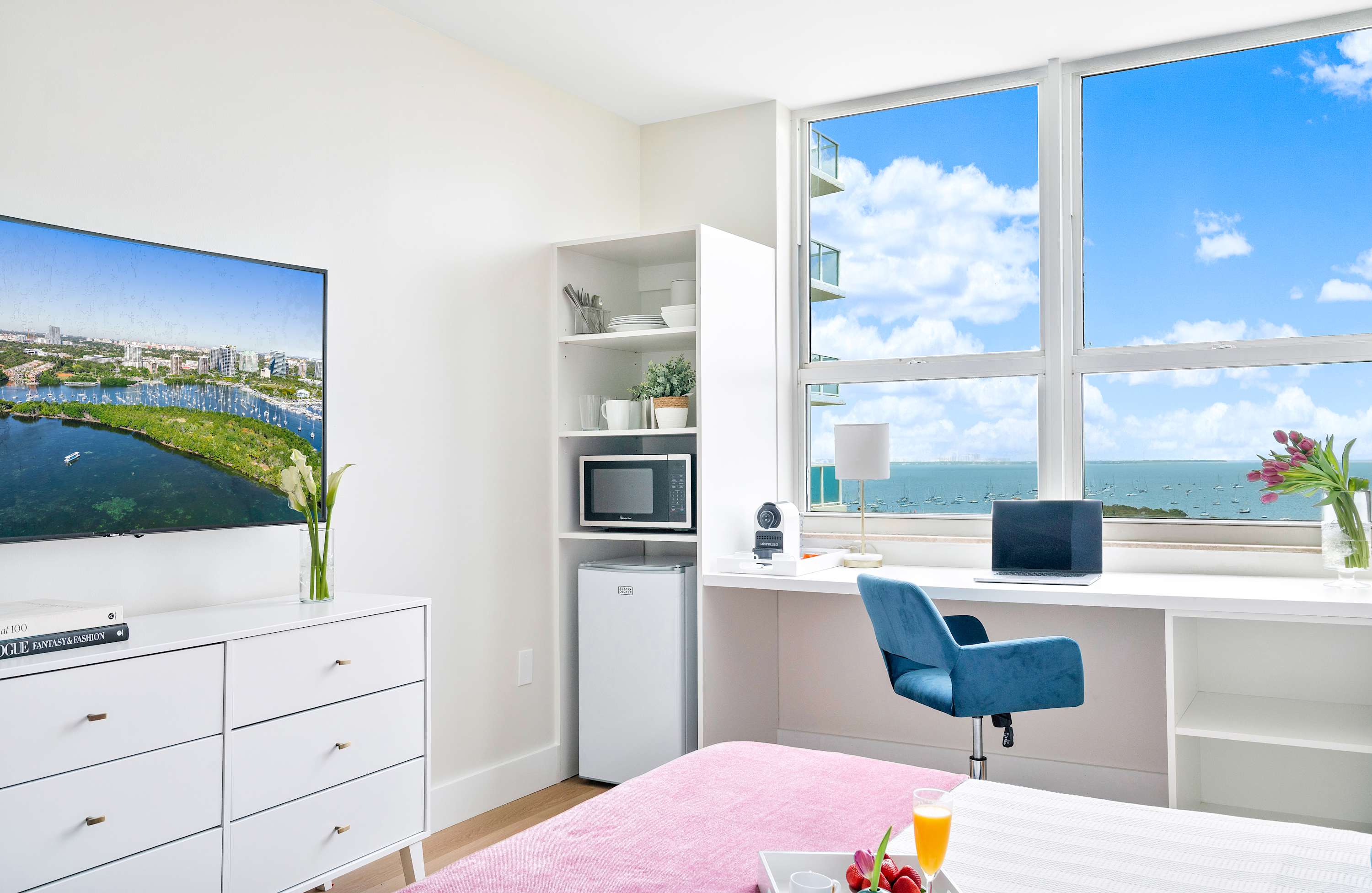 Modern bedroom featuring a white dresser with a flat-screen TV, a mini-fridge, microwave, and a built-in desk with an office chair, all with a stunning ocean view.