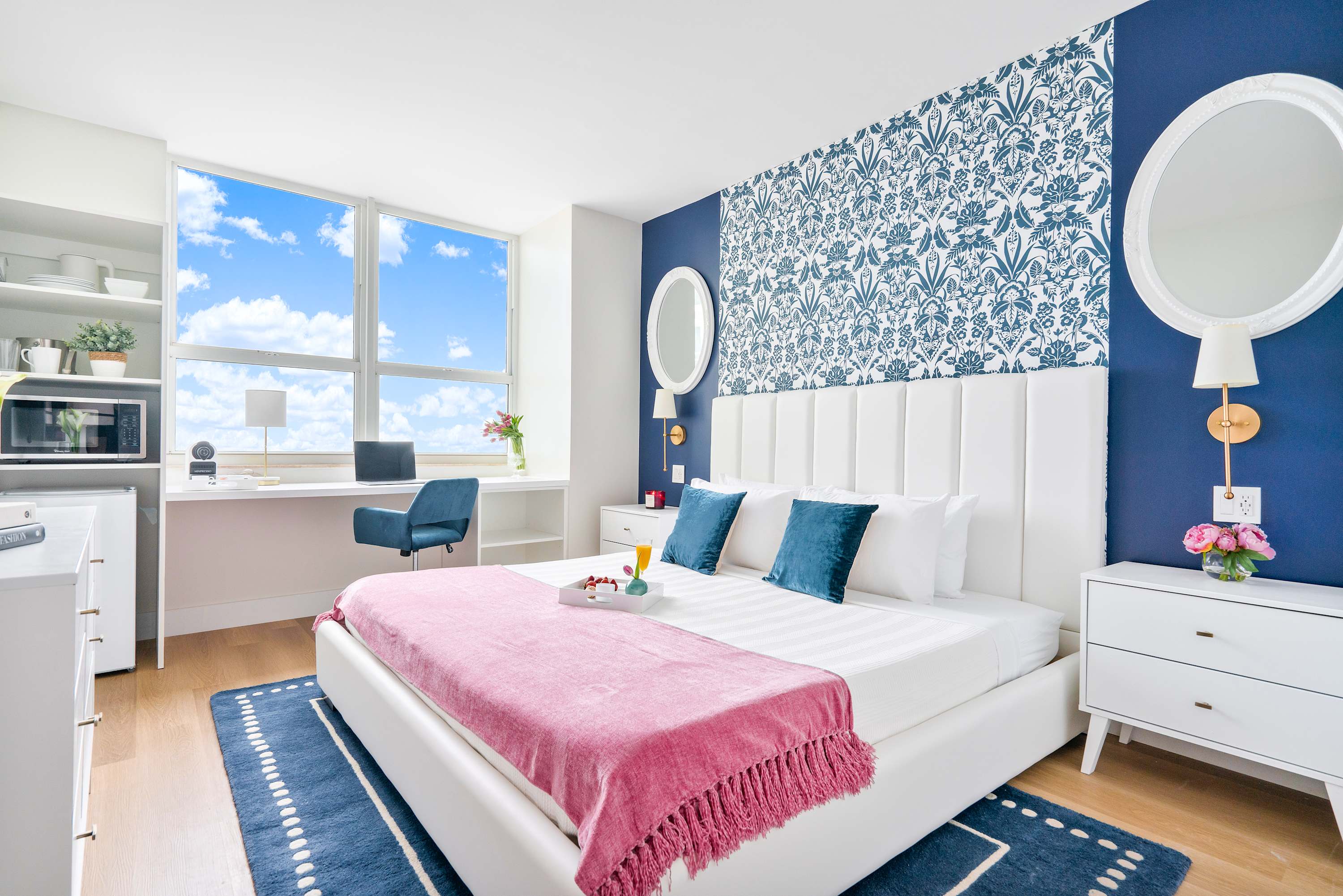 Bright bedroom with a white king bed, a blue and white patterned accent wall, and a dedicated workspace by a large window offering ocean views.
