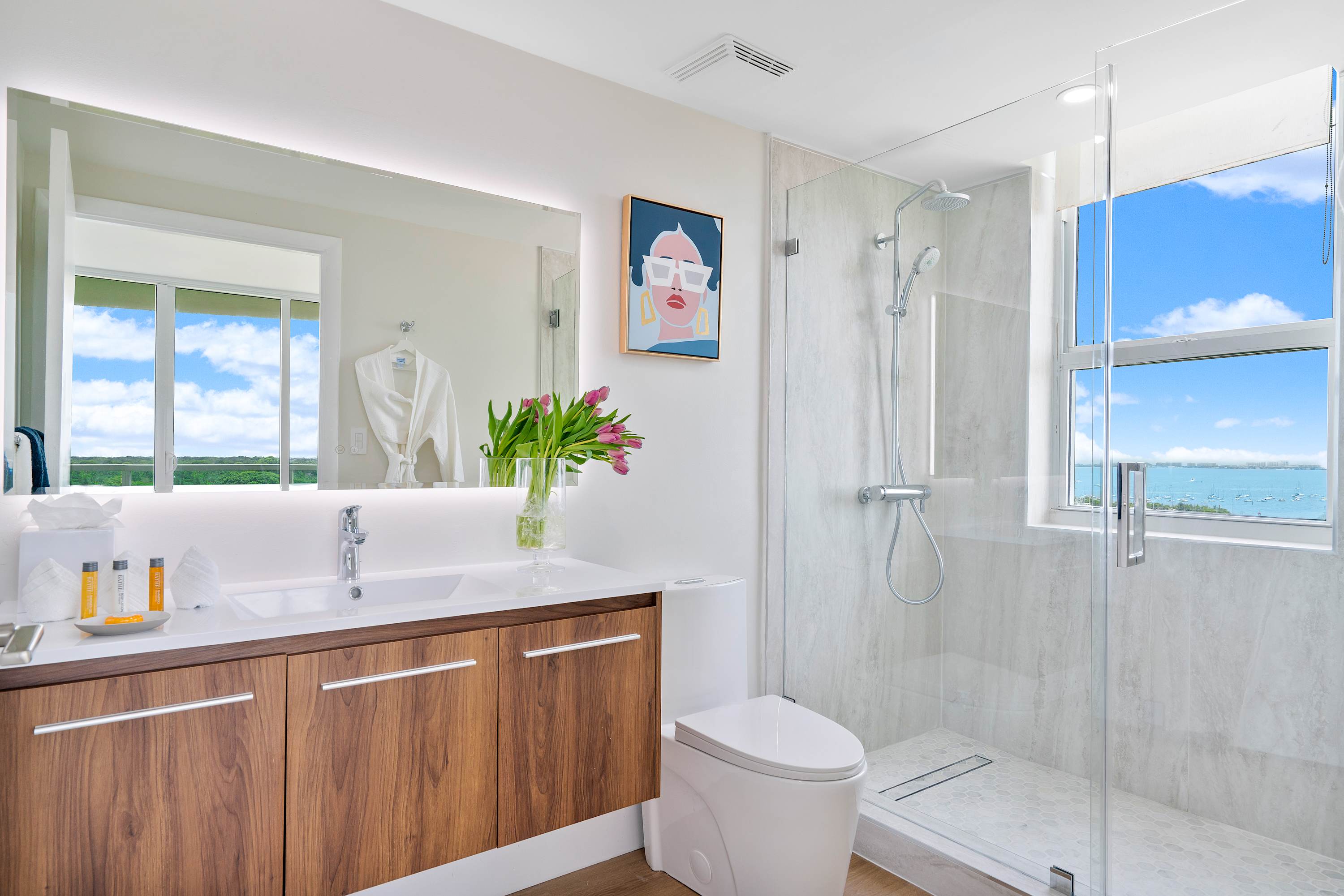 Contemporary bathroom with a wooden vanity, white countertop, and a walk-in glass shower. A large window in the shower offers expansive views of a bay with boats and a bright sky.