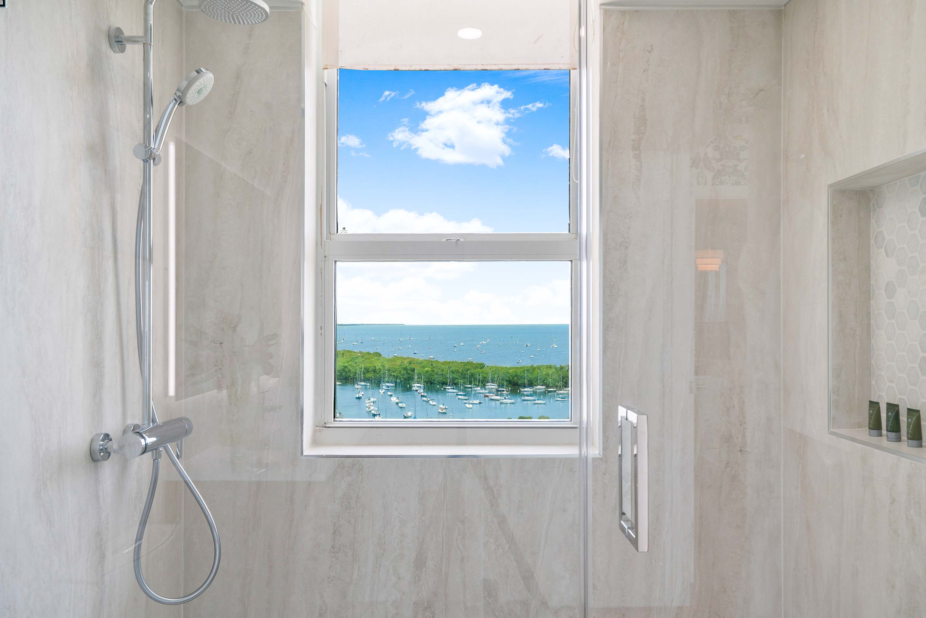 Contemporary walk-in shower with light-colored tiled walls, a rain shower head, and a large window offering panoramic views of a bay with boats and lush greenery.
