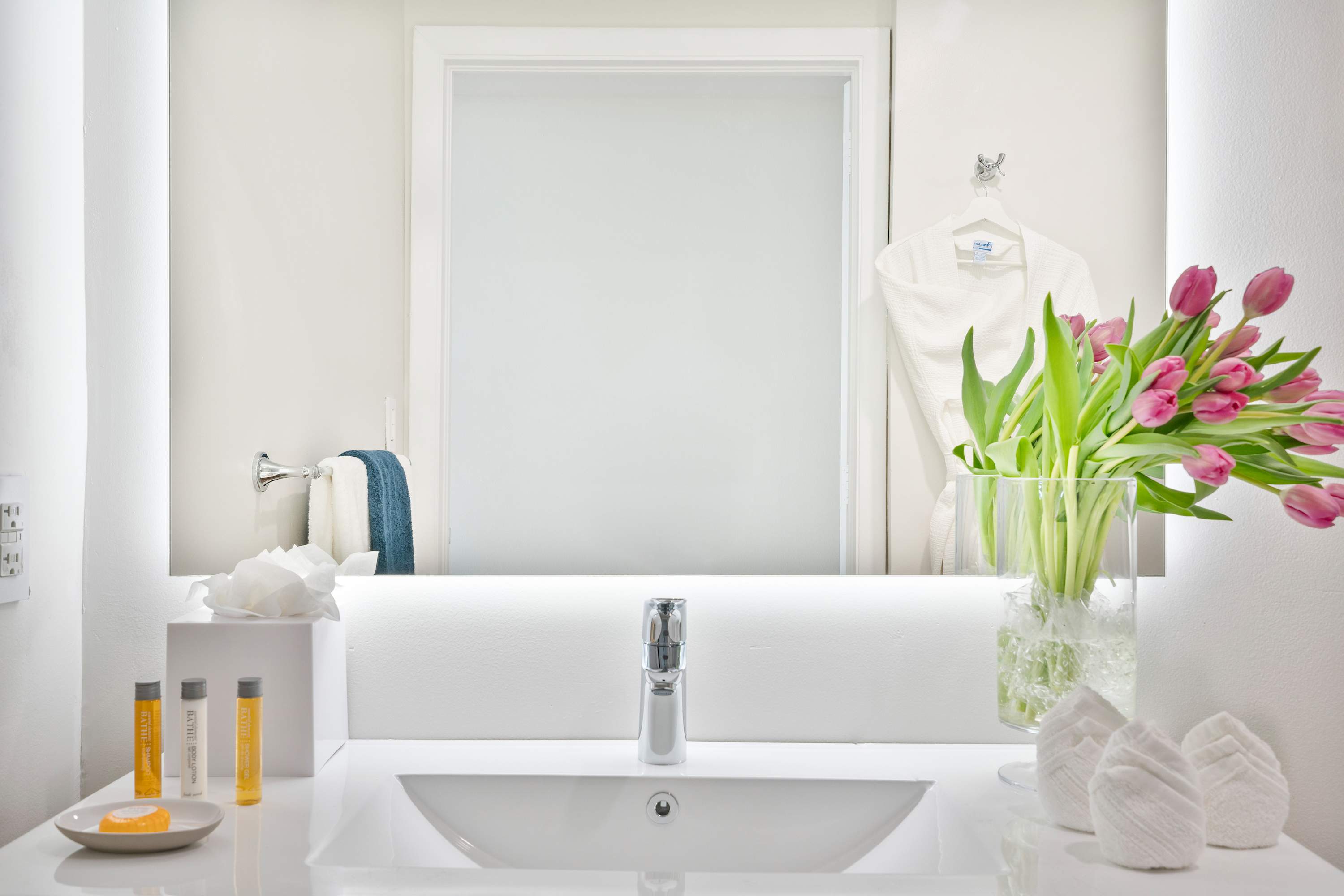 Modern white bathroom vanity with a rectangular sink, chrome faucet, and a large mirror reflecting a frosted door. Amenities like toiletries, a white bathrobe, and a vase of pink tulips are displayed.