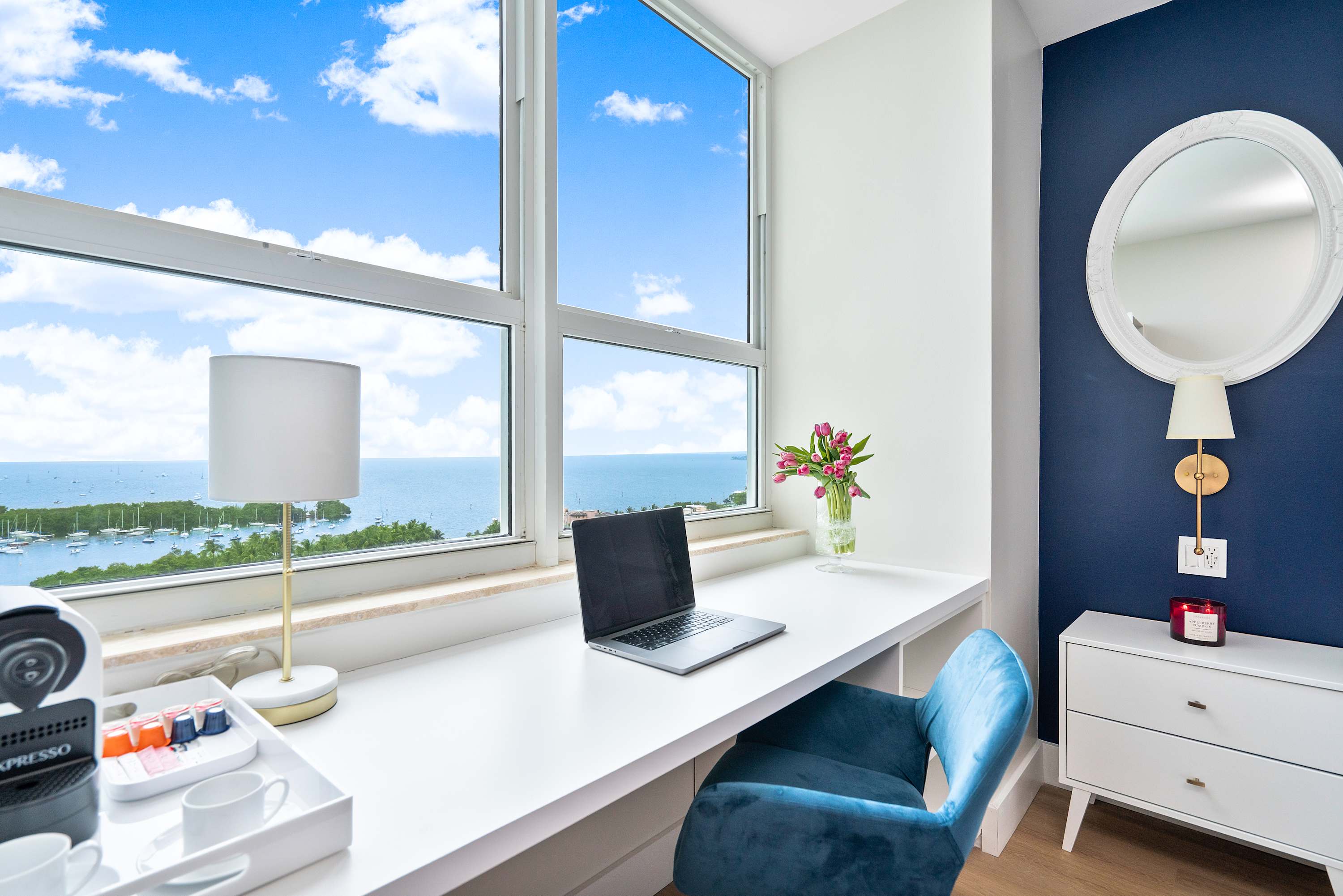 Dedicated workspace within the bedroom, featuring a white built-in desk and a blue velvet office chair, positioned in front of large windows offering panoramic ocean and marina views.