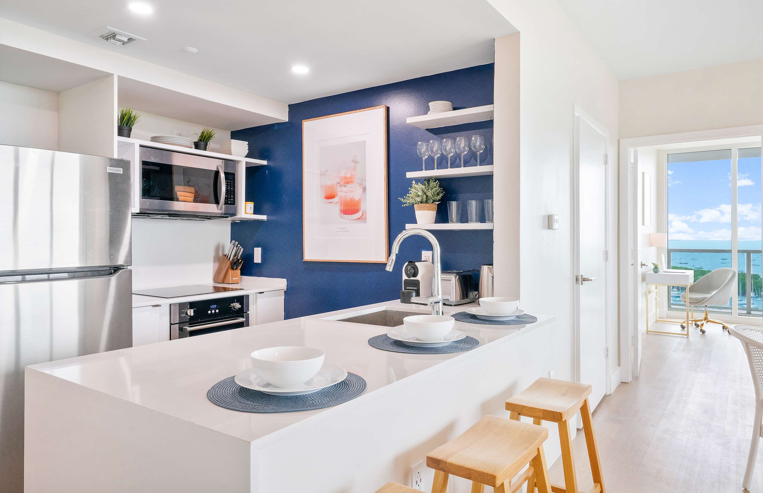 Contemporary kitchen featuring a white countertop island with bar seating, stainless steel appliances, open shelving with decor, and a vibrant blue accent wall.