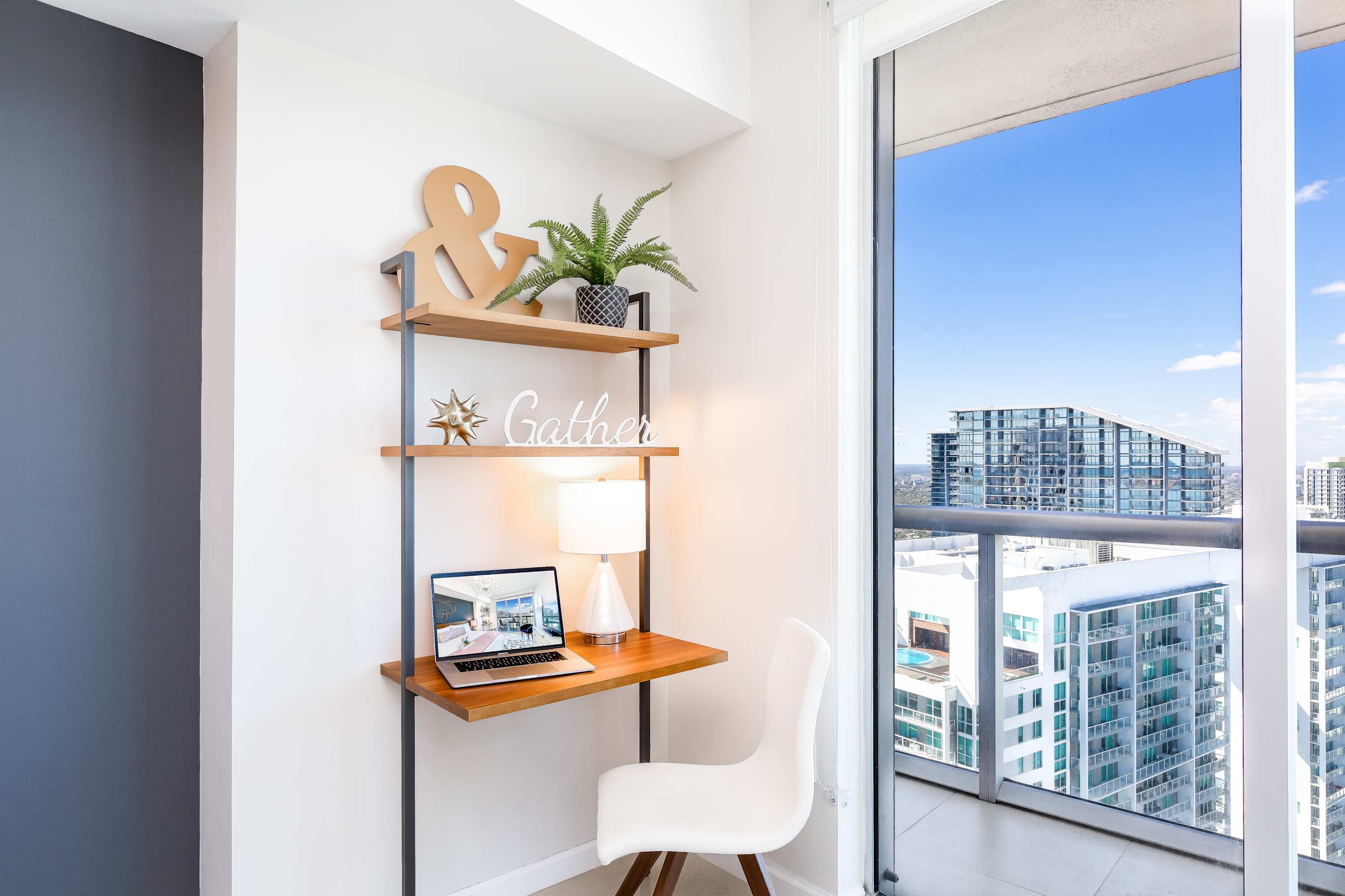 Compact home office nook with a wooden desk and shelving, positioned next to a glass sliding door leading to a balcony with expansive city views.