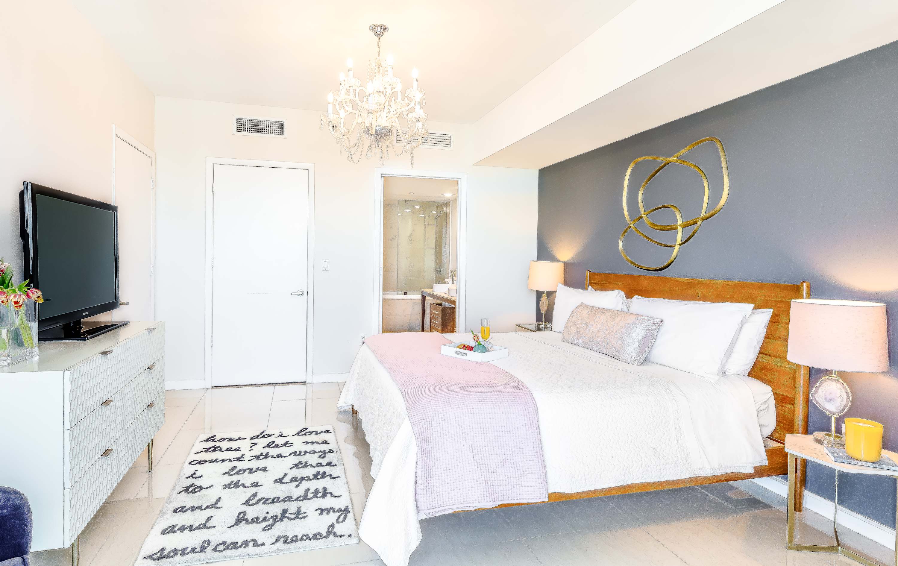 The spacious bedroom showcases a large bed with a wooden headboard and a grey accent wall, complemented by a luxurious chandelier and a white dresser with a TV. A unique rug with text adds character, while a glimpse of the en-suite bathroom is visible through an open doorway.