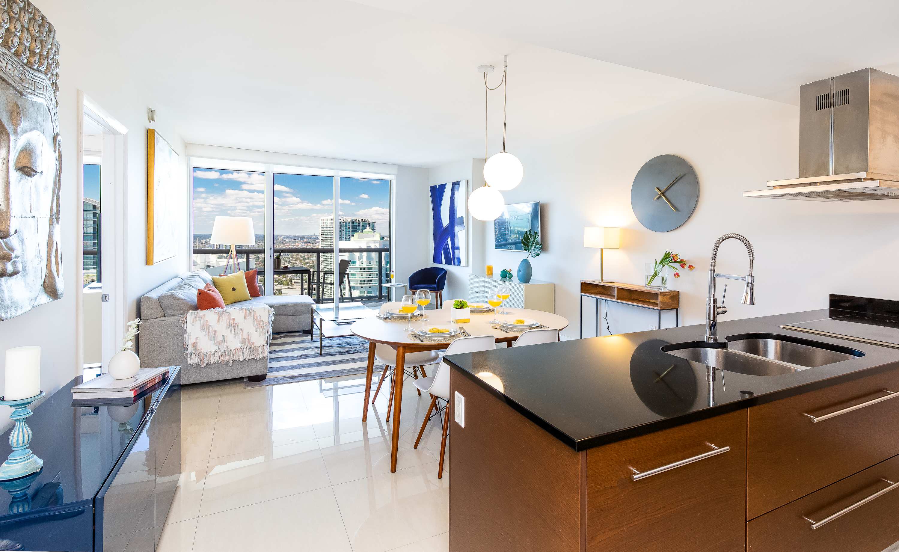 An expansive open-concept living space seamlessly connects a modern kitchen with dark countertops and wooden cabinetry to a dining area with a round white table and a bright living room. Large windows frame a stunning city view, enhancing the contemporary and airy ambiance.