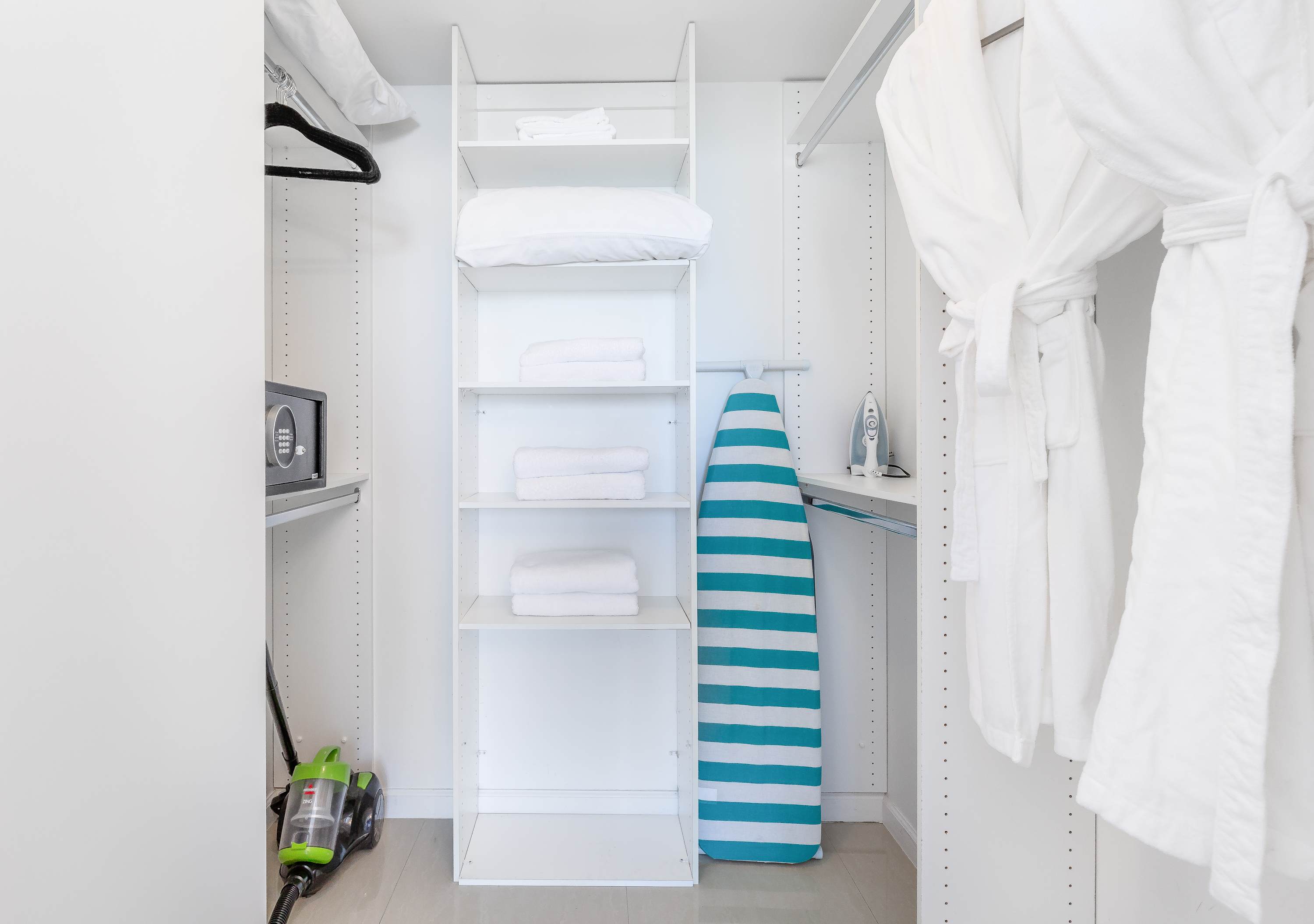 Organized walk-in closet with white shelving, a safe, an ironing board, and hanging white robes, providing ample storage space.
