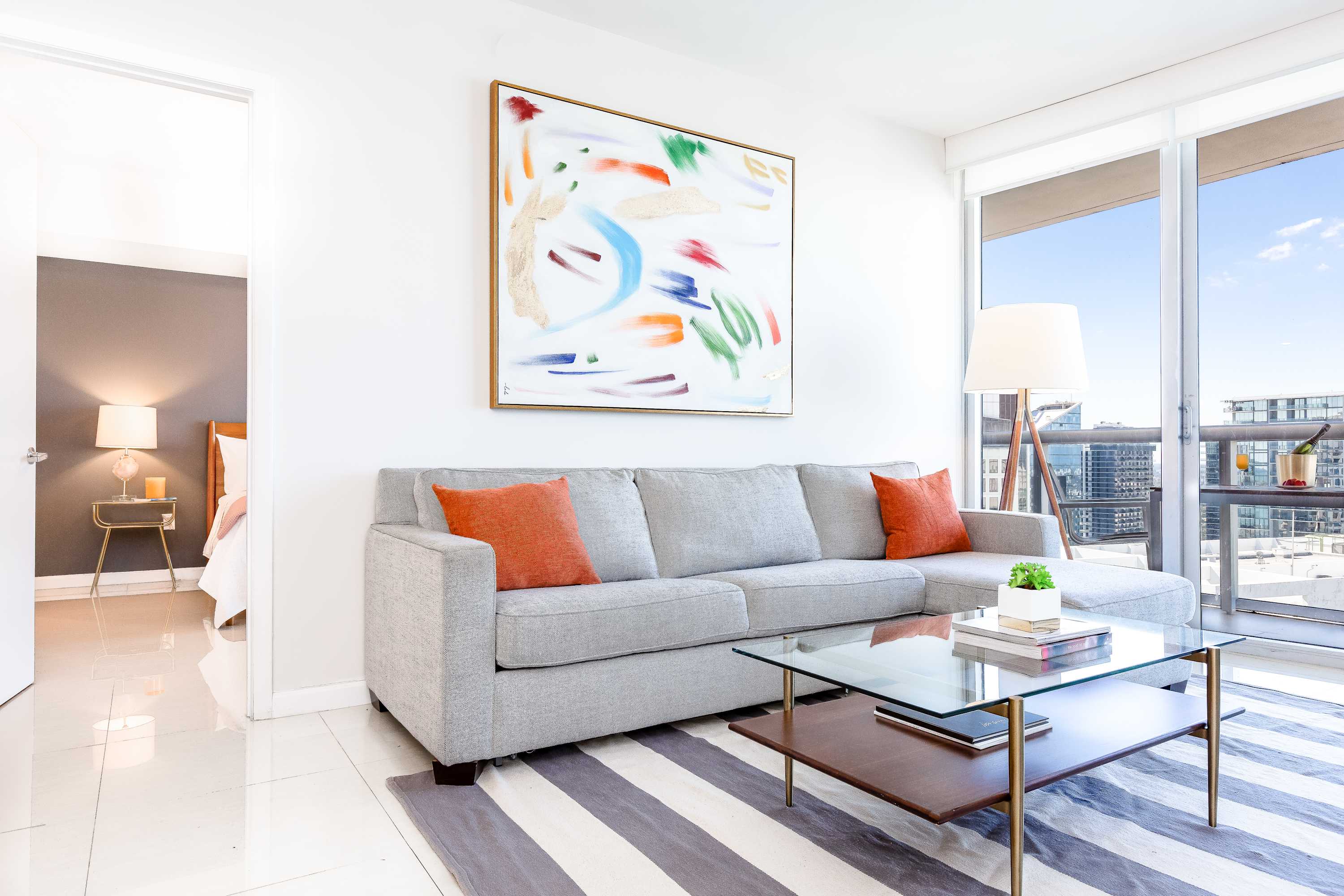 A modern living room features a comfortable grey sectional sofa adorned with orange throw pillows, facing large windows that offer city views. A glass-top coffee table sits on a striped rug, and a colorful abstract painting hangs above the sofa, adding a vibrant touch to the bright space.