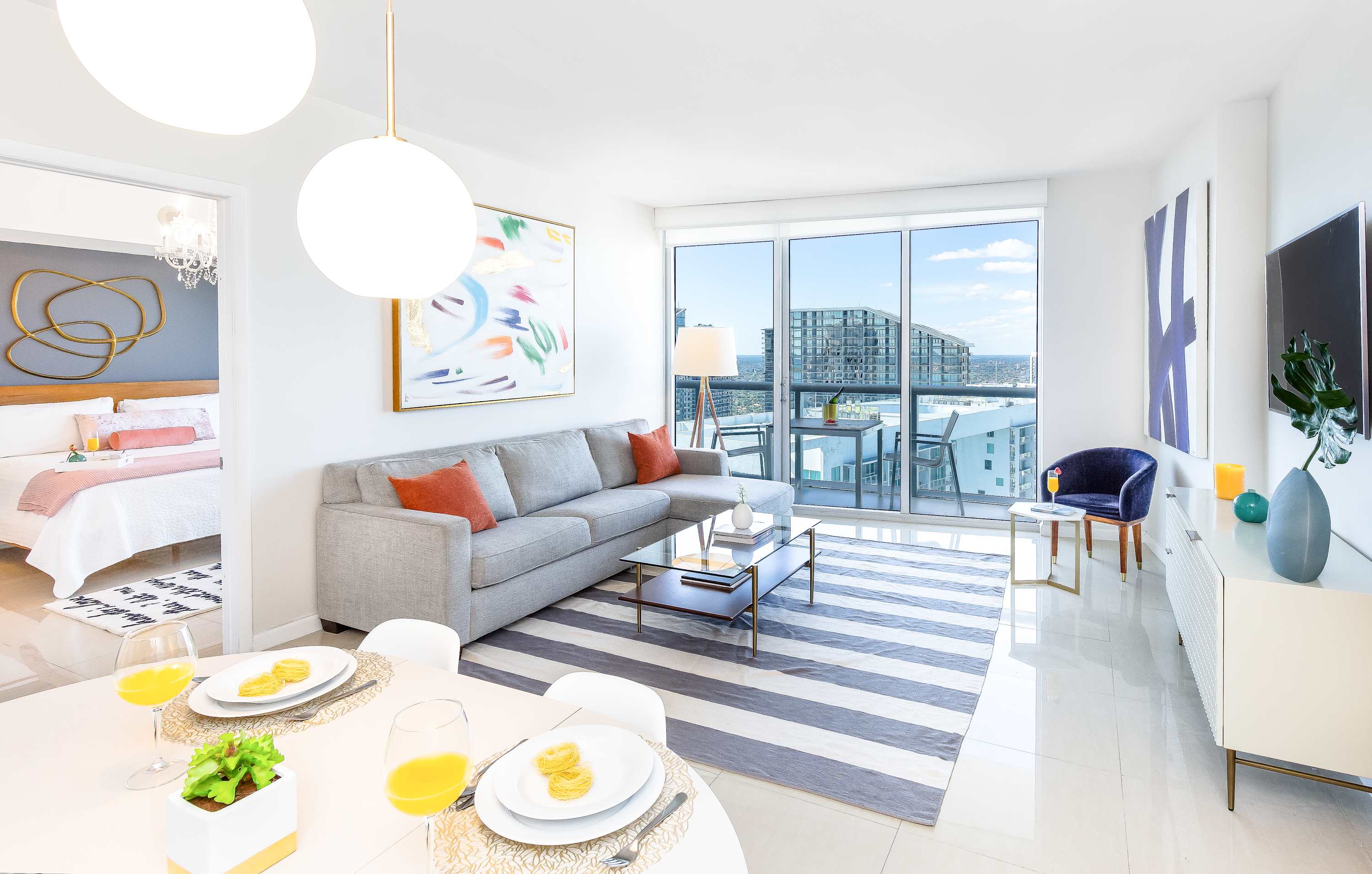 Bright, open-concept living and dining area with a grey sectional sofa, striped rug, modern dining table, and large windows offering city views.