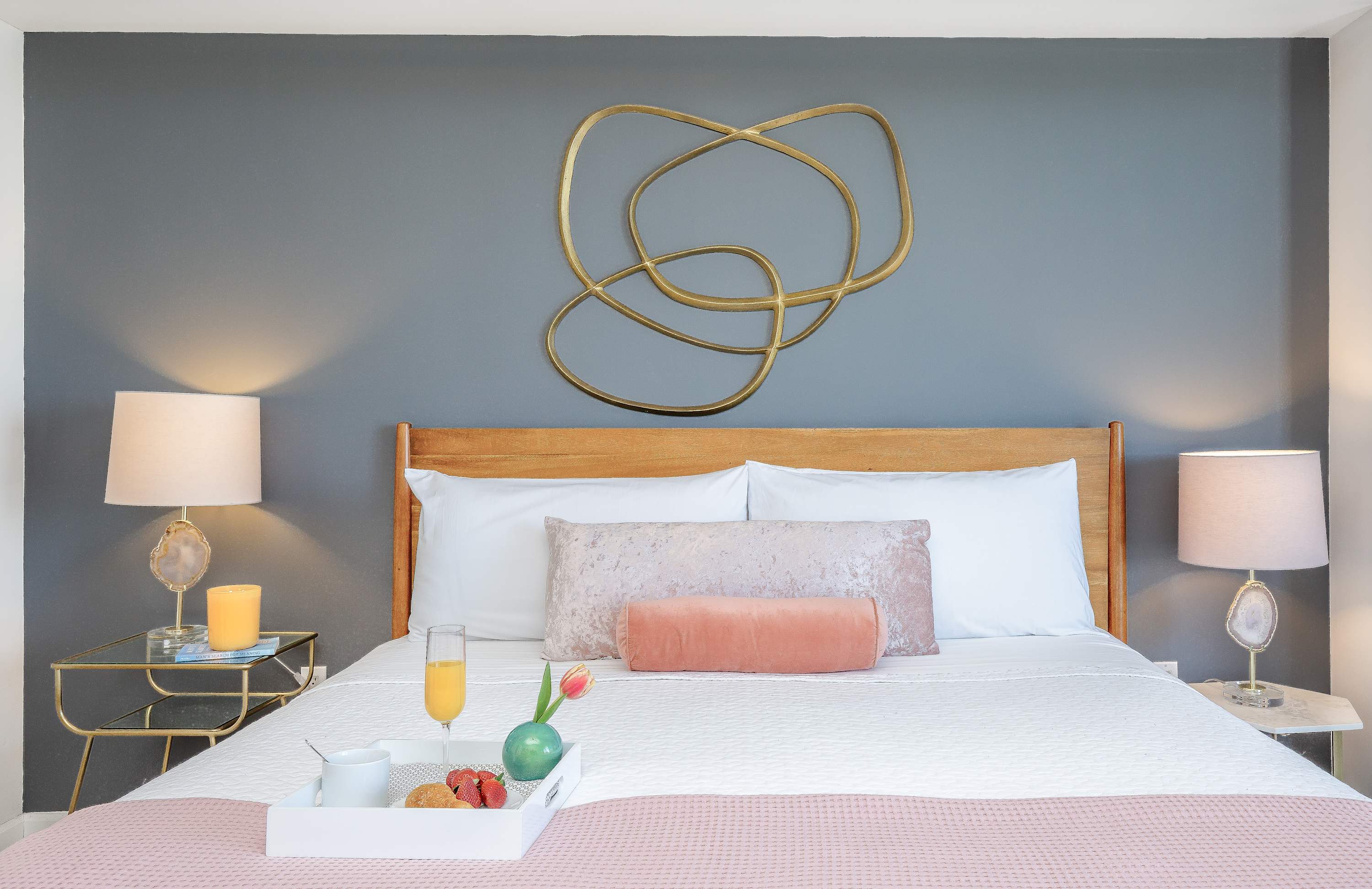 Cozy bedroom with a wooden headboard, white bedding, pink accent pillows, and two nightstands with lamps, featuring a gold abstract wall art.