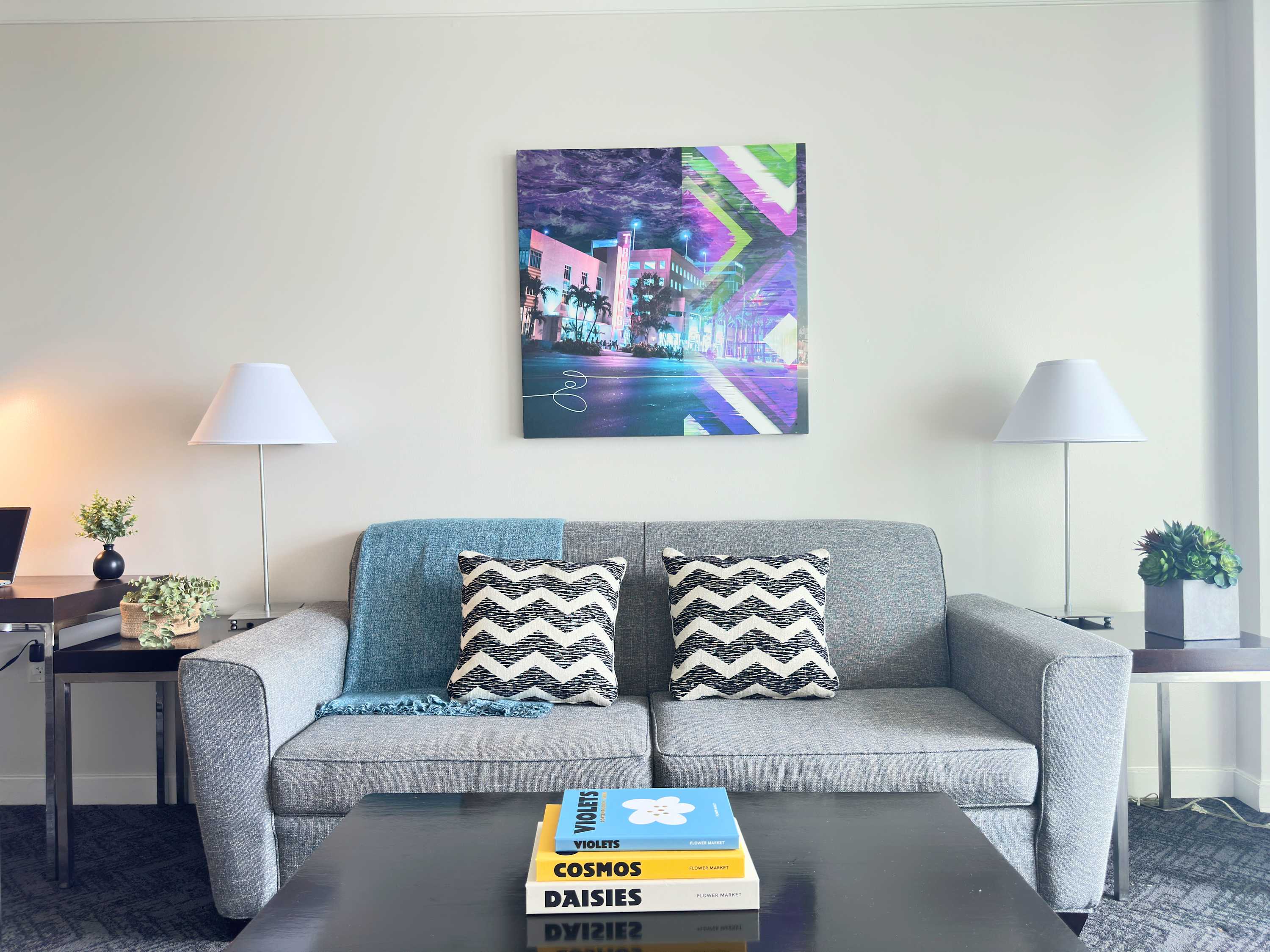 Cozy living room featuring a gray sofa with patterned throw pillows, a dark coffee table with books, and a vibrant abstract art piece on a light wall.