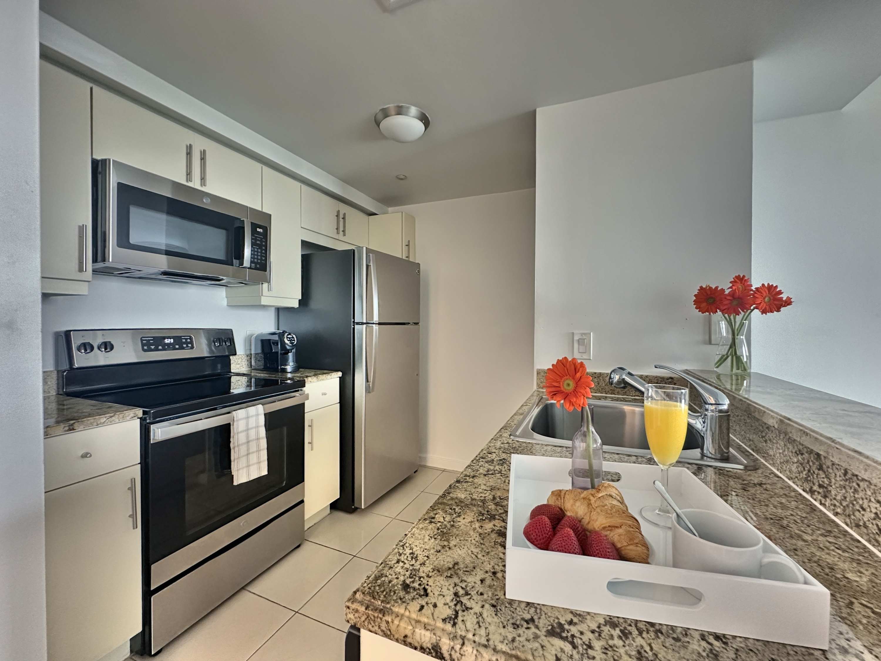 Modern kitchen equipped with stainless steel appliances, light wood cabinetry, granite countertops, and a breakfast tray with orange juice and pastries.