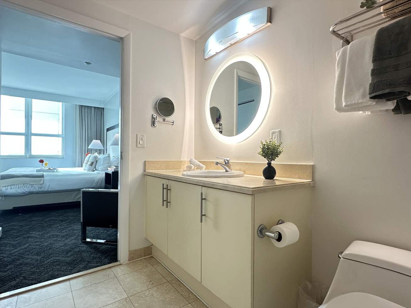 Modern half bathroom with a light-colored vanity, a circular backlit mirror, and a toilet, offering a glimpse into the adjacent bedroom.
