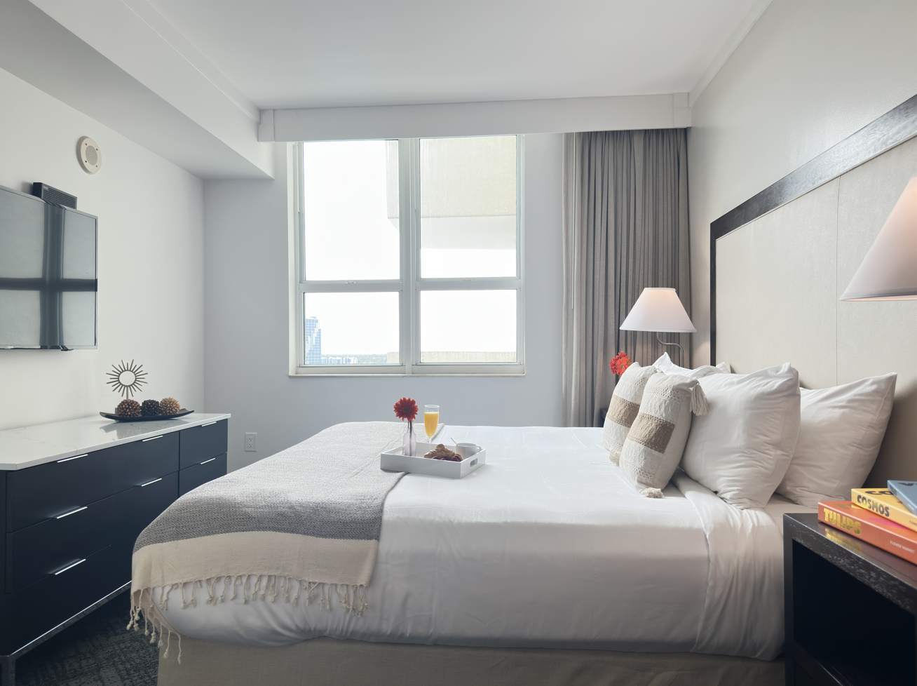 Bright bedroom featuring a king-sized bed with white bedding and a light gray throw, a large window with city views, and a dark dresser with a wall-mounted TV.