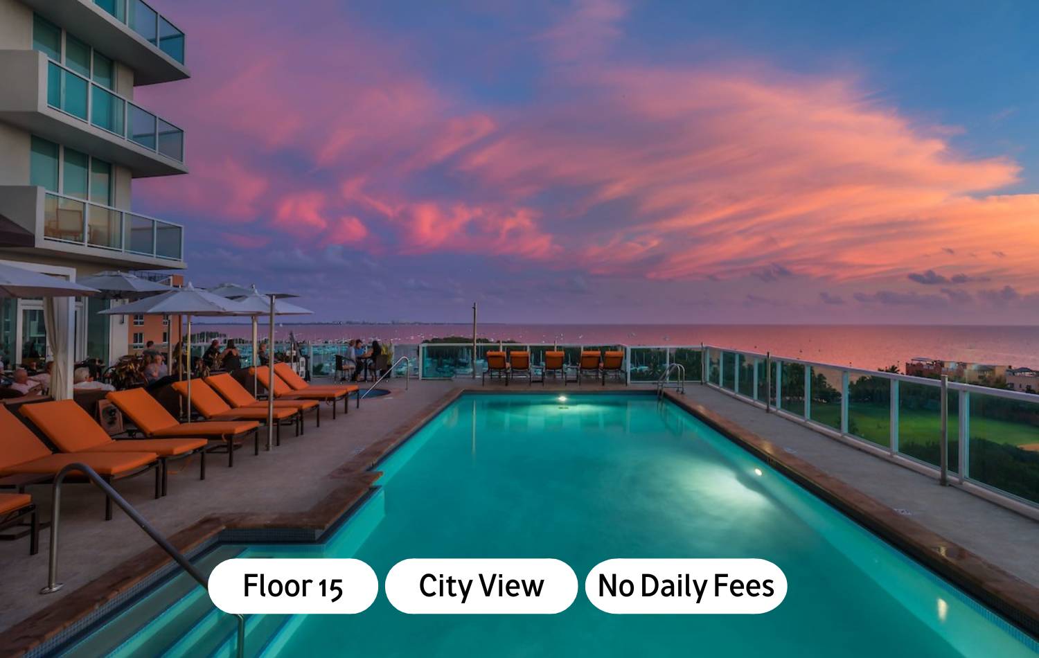 Rooftop swimming pool with orange lounge chairs, offering panoramic city and ocean views under a vibrant sunset sky, adjacent to a modern building facade.